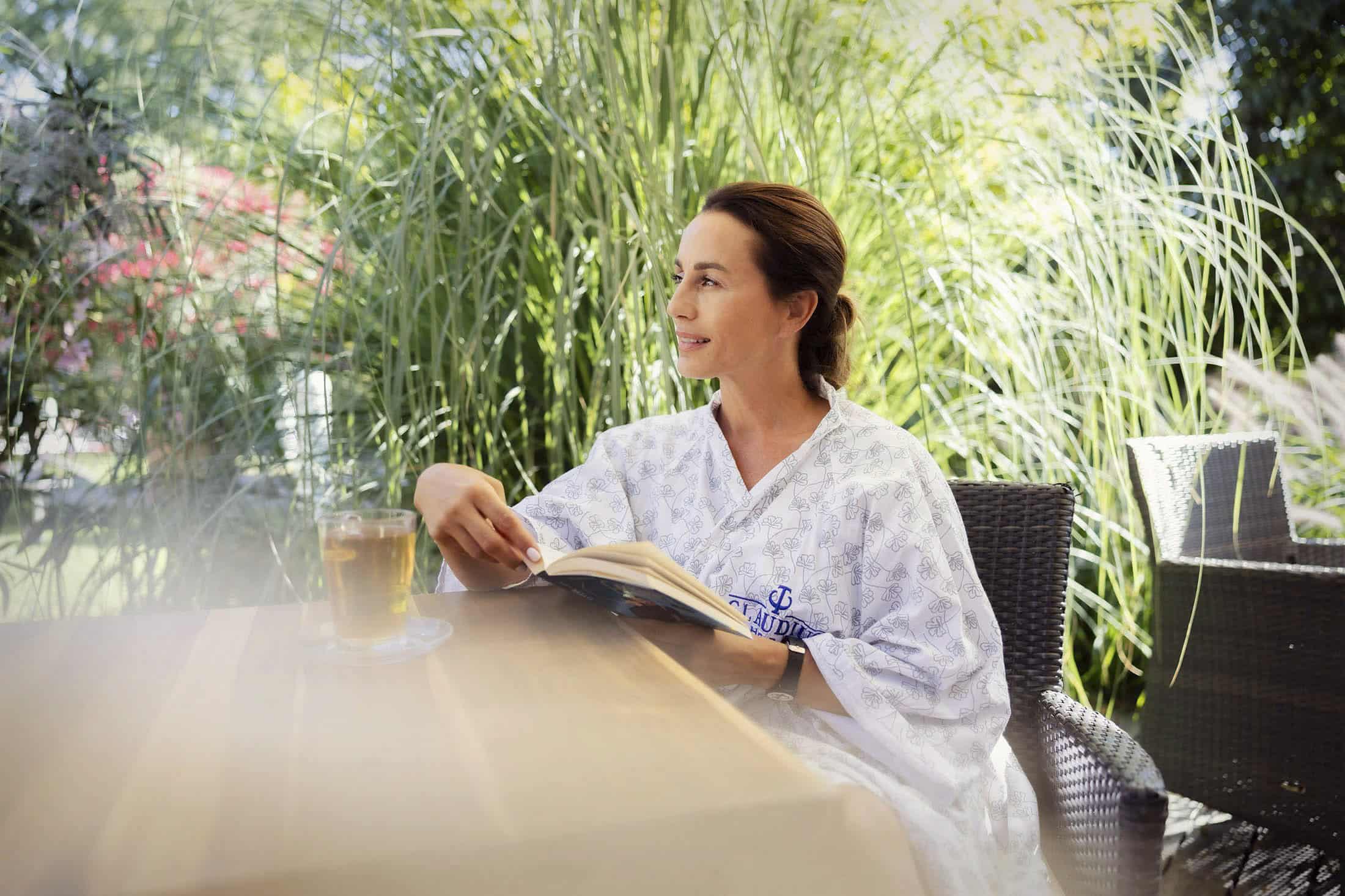 Eine Person sitzt an einem Tisch im Freien, umgeben von hohem Gras. Sie trägt einen leichten Bademantel und blickt zur Seite. Sie hält ein offenes Buch in der Hand und auf dem Tisch steht eine Tasse Tee. Sonnenlicht fällt durch das Grün und schafft eine friedliche Atmosphäre. © Fotografie Tomas Rodriguez