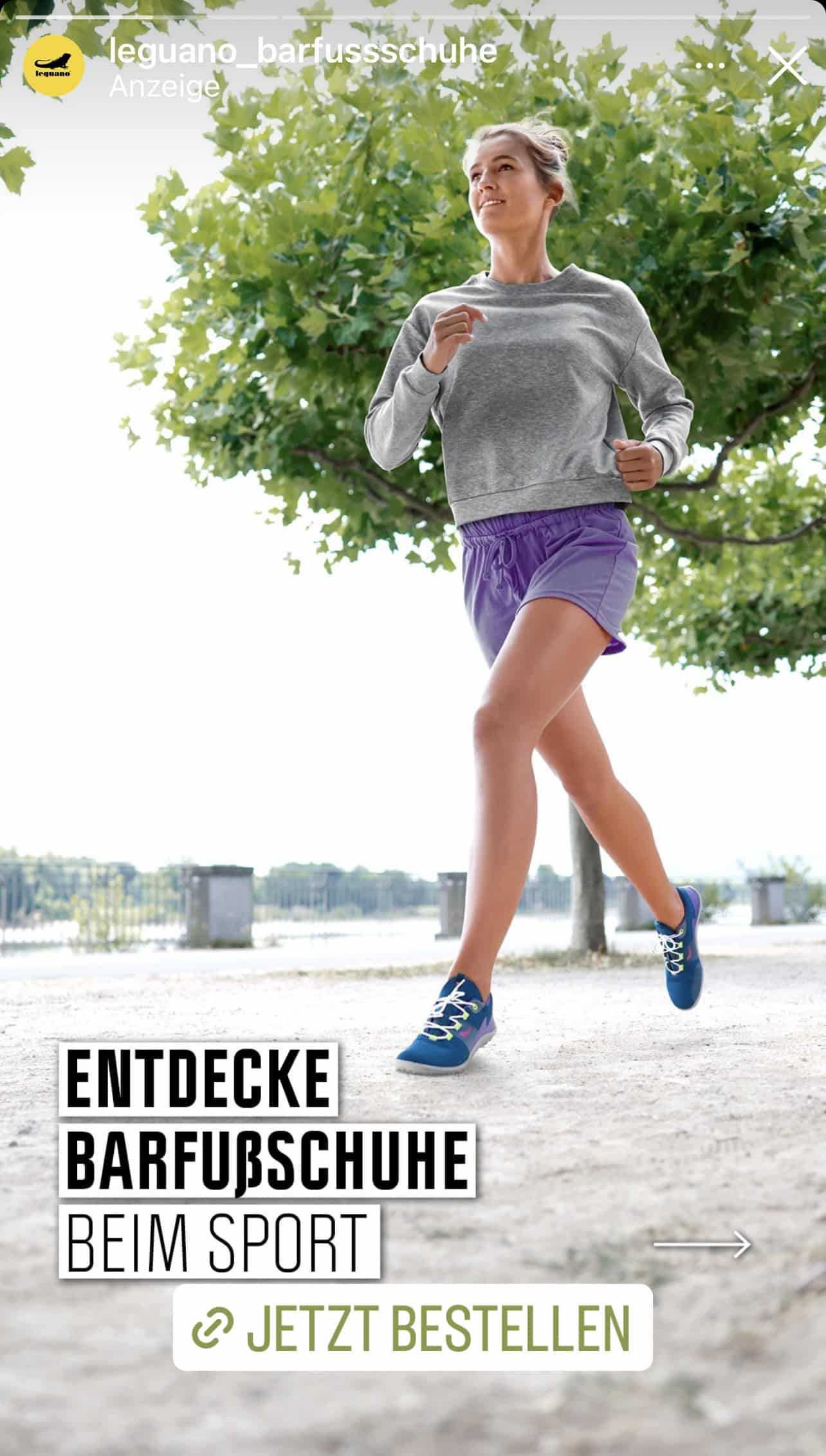 Eine Frau joggt auf einem gepflasterten Weg und trägt ein graues Sweatshirt, lila Shorts und blaue Schuhe. Sie steht neben einem grünen Baum, in der Ferne sind ein Geländer und eine Stadtlandschaft zu sehen. Texteinblendung: „ENTDECKE BARFUSSSCHUHE BEIM SPORT – JETZT BESTELLEN. © Fotografie Tomas Rodriguez