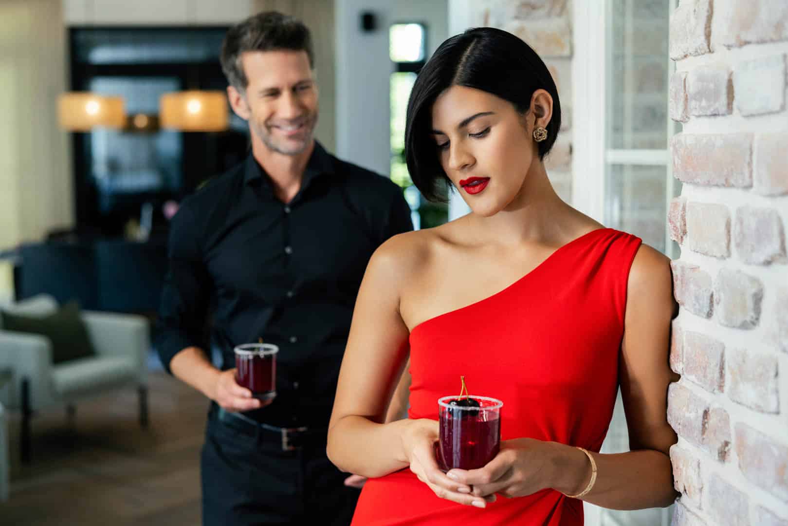 Eine Frau in einem roten One-Shoulder-Kleid hält ein Getränk in der Hand, lehnt an einer Backsteinmauer und blickt nach unten. Im Hintergrund steht ein Mann in einem schwarzen Hemd, hält ein ähnliches Getränk in der Hand und lächelt sie an. Die Szenerie ist ein elegant eingerichteter Raum. © Fotografie Tomas Rodriguez