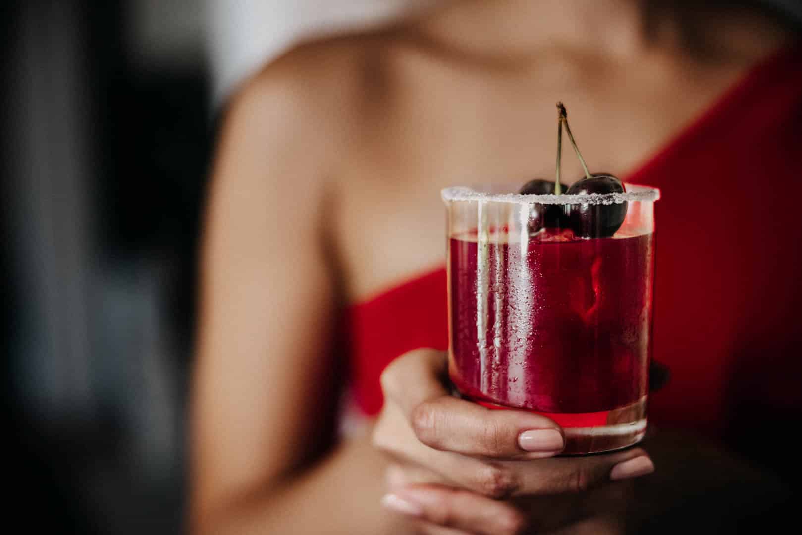 Eine Person hält ein Glas mit einem roten Getränk, garniert mit zwei Kirschen, in einer Umgebung mit dezenter Beleuchtung. Die Person trägt ein rotes, schulterfreies Outfit und das Glas hat einen Salz- oder Zuckerrand. © Fotografie Tomas Rodriguez