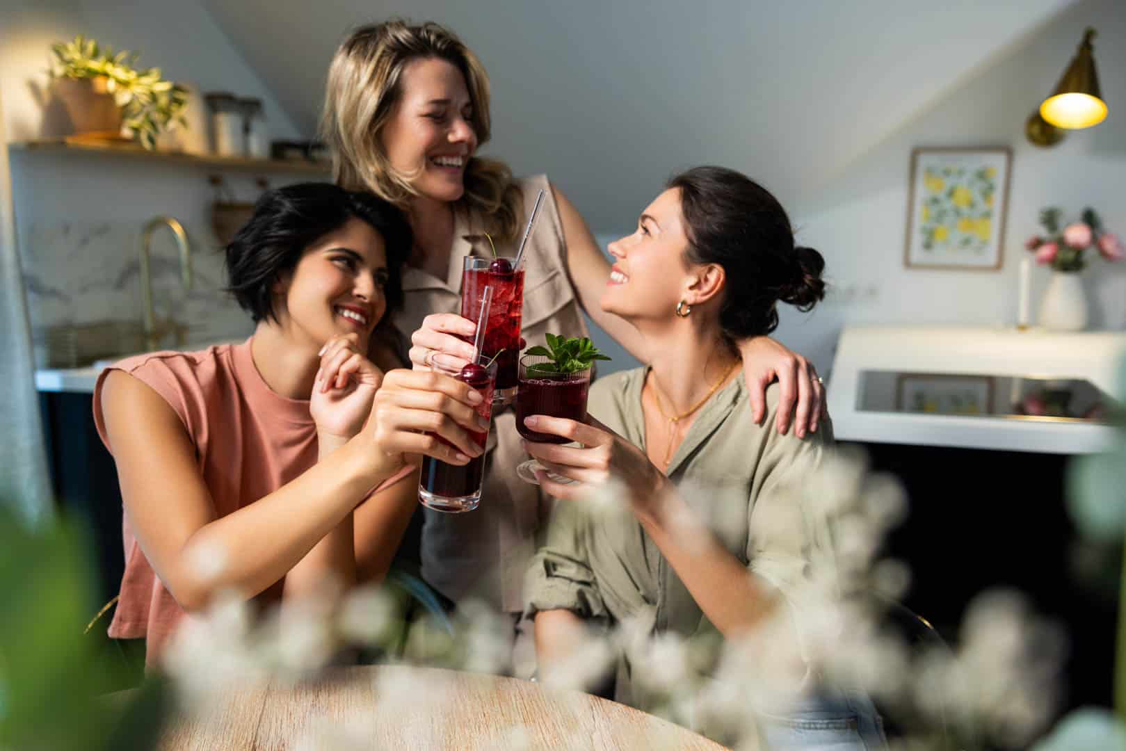 Drei Frauen lächeln und stoßen mit Getränken in einer modernen Küche an. Sie sitzen um einen Tisch herum, im Vordergrund ist eine verschwommene Pflanze zu sehen. Die Atmosphäre ist fröhlich und locker. © Fotografie Tomas Rodriguez