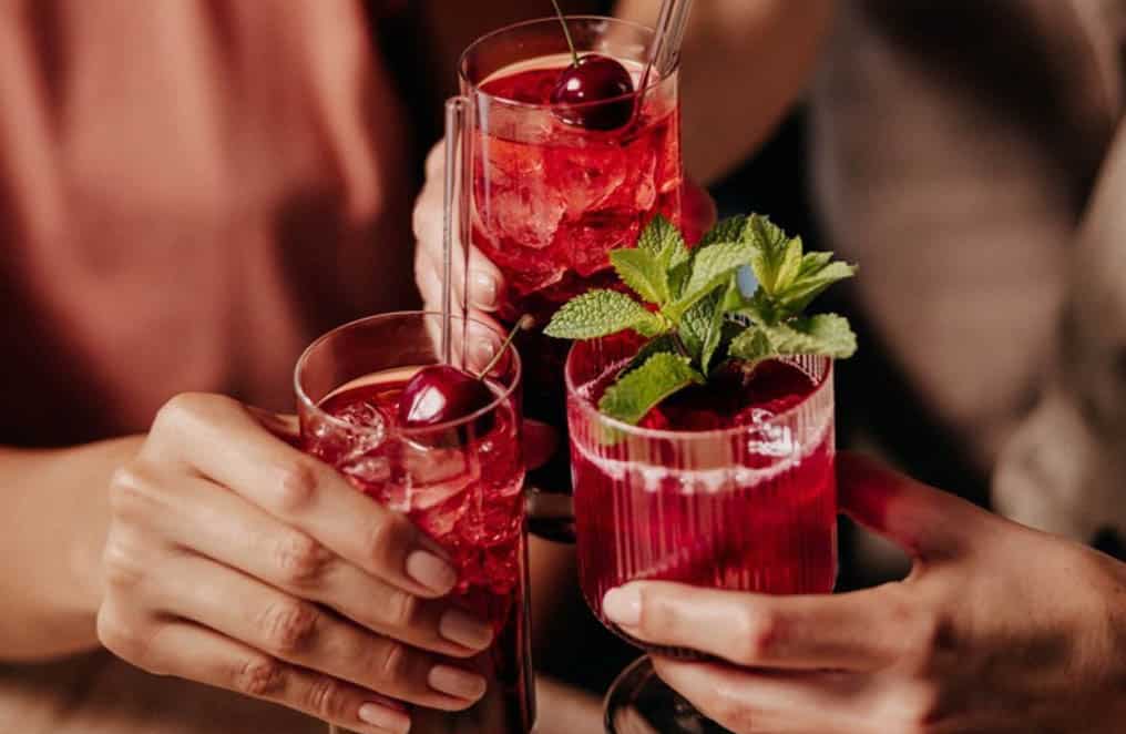 Drei Hände halten Gläser mit roten Cocktails. Die Getränke sind mit Kirschen und Minzblättern garniert. Die Szene vermittelt ein Gefühl von Feier oder Zusammensein. © Fotografie Tomas Rodriguez