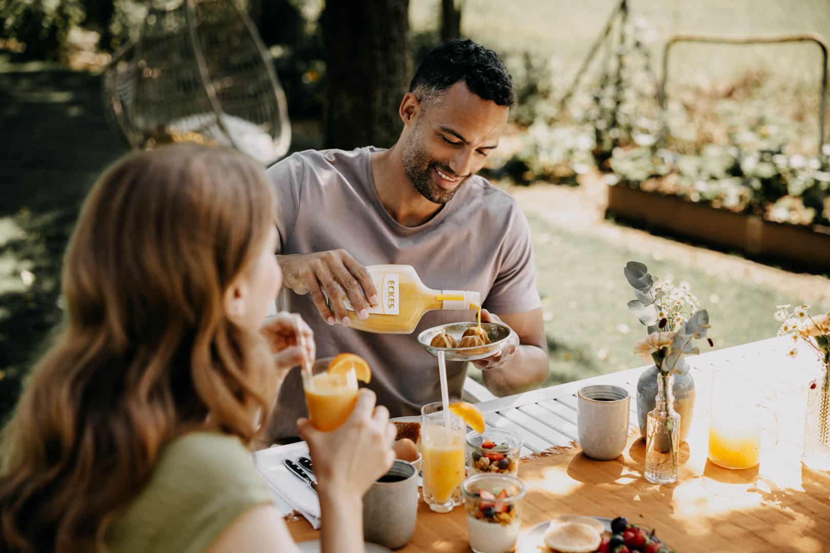 An einem Tisch im Freien gießt ein Mann Orangensaft in das Glas einer Frau. Sie genießen einen Brunch mit Obsttellern und Gebäck in einer Gartenumgebung. Sonnenlicht fällt durch die Bäume und schafft eine entspannte und fröhliche Atmosphäre. © Fotografie Tomas Rodriguez