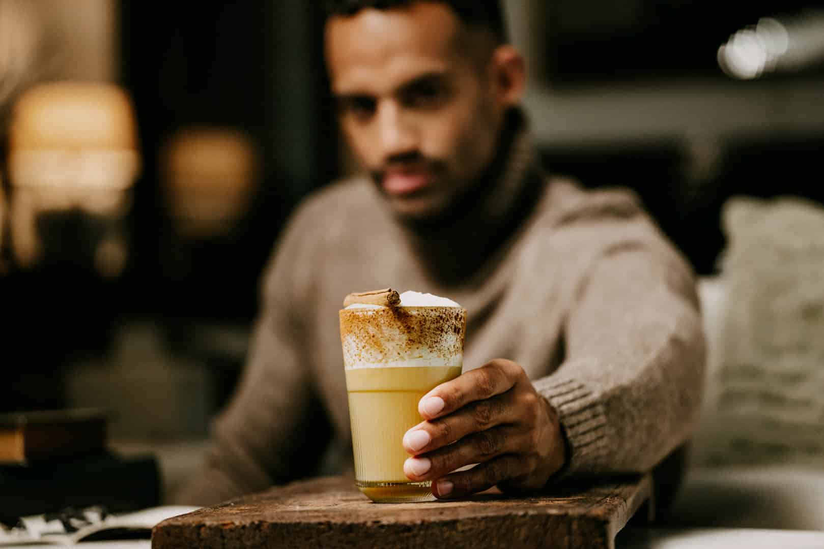 Eine Person in einem gemütlichen Pullover hält ein Glas Kaffee mit Schlagsahne und Zimt hoch, das auf einer Holzoberfläche steht. Der Hintergrund ist sanft verschwommen und schafft eine warme, einladende Atmosphäre. © Fotografie Tomas Rodriguez