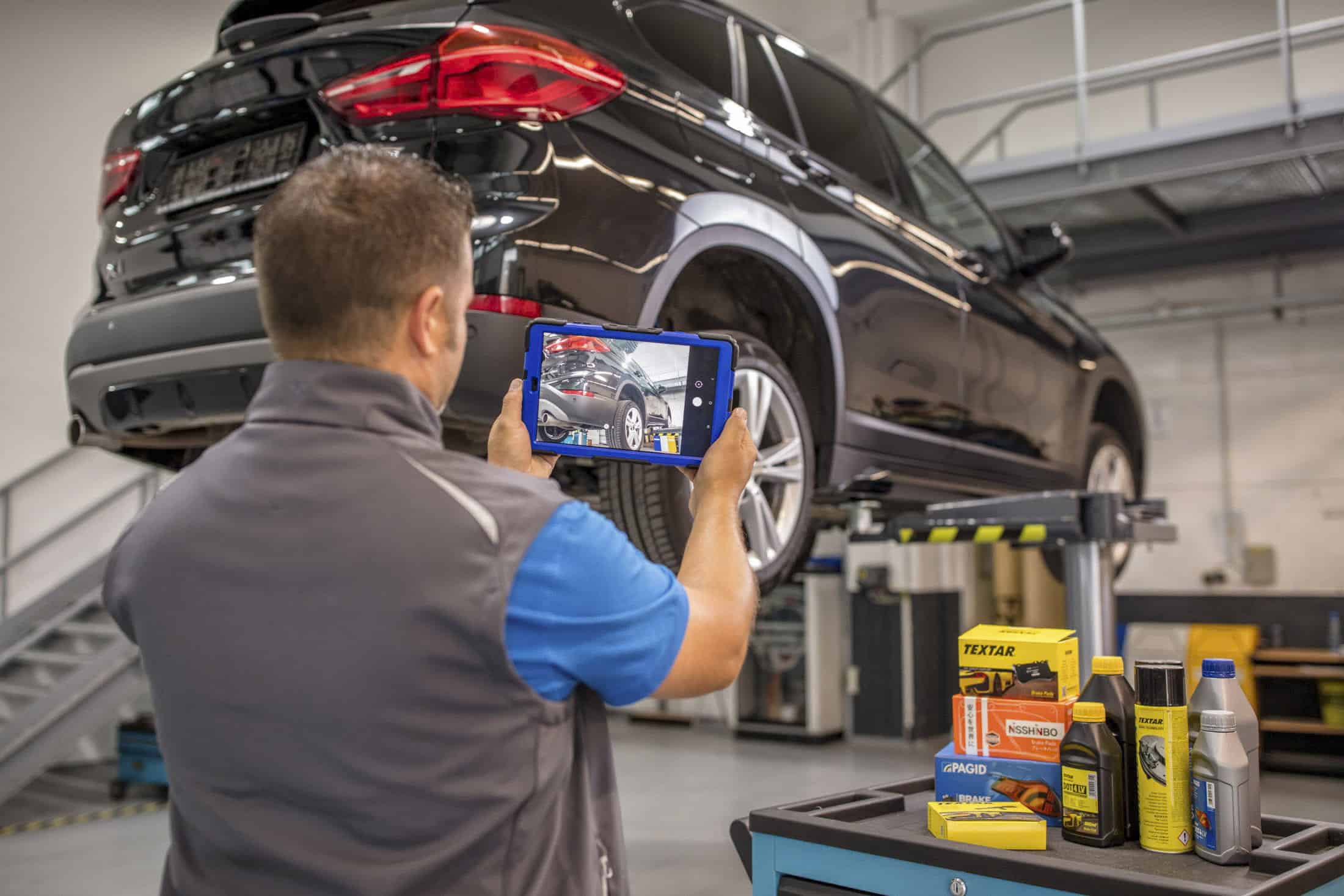Ein Mechaniker überprüft mit einem Tablet einen schwarzen SUV, der in einer Garage auf einer Hebebühne steht. Auf einem daneben stehenden Wagen sind verschiedene Automobilprodukte ausgestellt, darunter Bremsbeläge und Motoröle. Die Werkstatt ist gut beleuchtet und hat graue Böden und Regale. © Fotografie Tomas Rodriguez