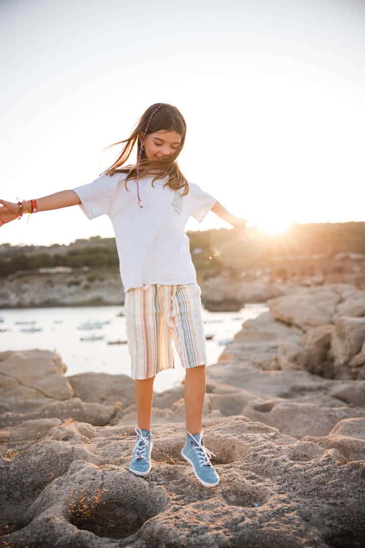 Ein junges Mädchen mit langen Haaren balanciert auf felsigem Gelände. Es trägt ein weißes Hemd, gestreifte Shorts und blaue Turnschuhe. Im Hintergrund geht die Sonne unter und beleuchtet ein ruhiges Gewässer und Boote in der Ferne. Sie sieht zufrieden und entspannt aus. © Fotografie Tomas Rodriguez