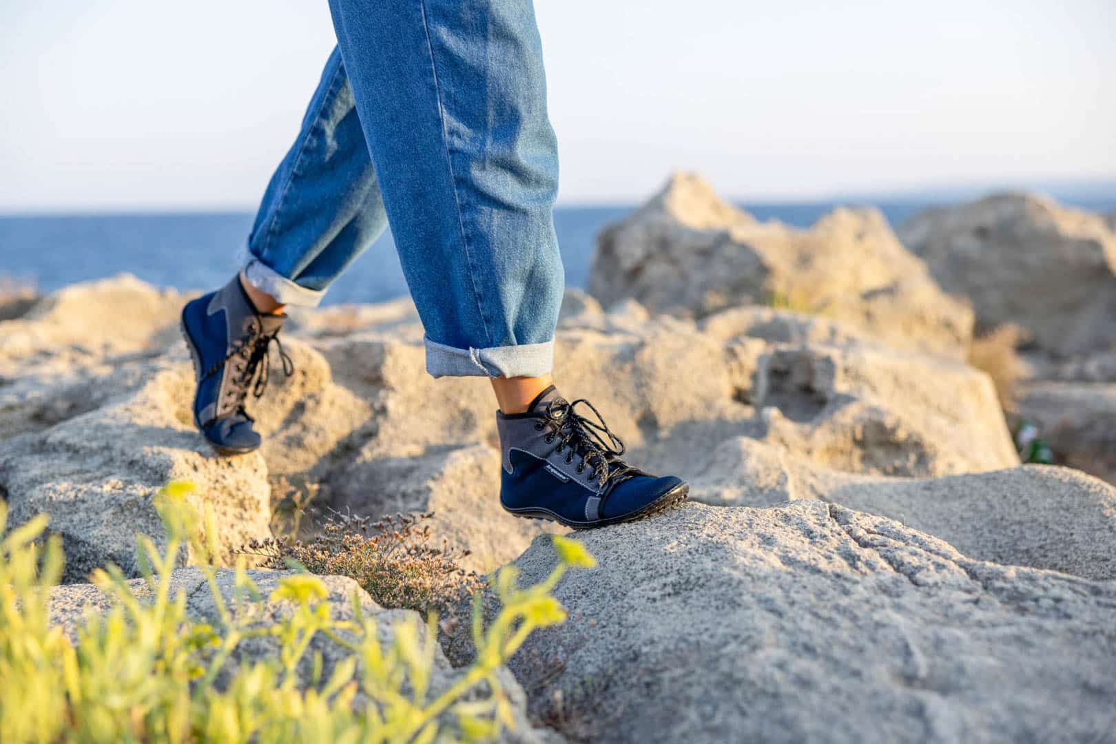 Eine Person in hochgekrempelten Bluejeans und marineblauen Wanderschuhen läuft über felsiges Gelände in der Nähe des Ozeans, mit Pflanzen im Vordergrund und dem Meer im Hintergrund unter einem klaren Himmel. © Fotografie Tomas Rodriguez