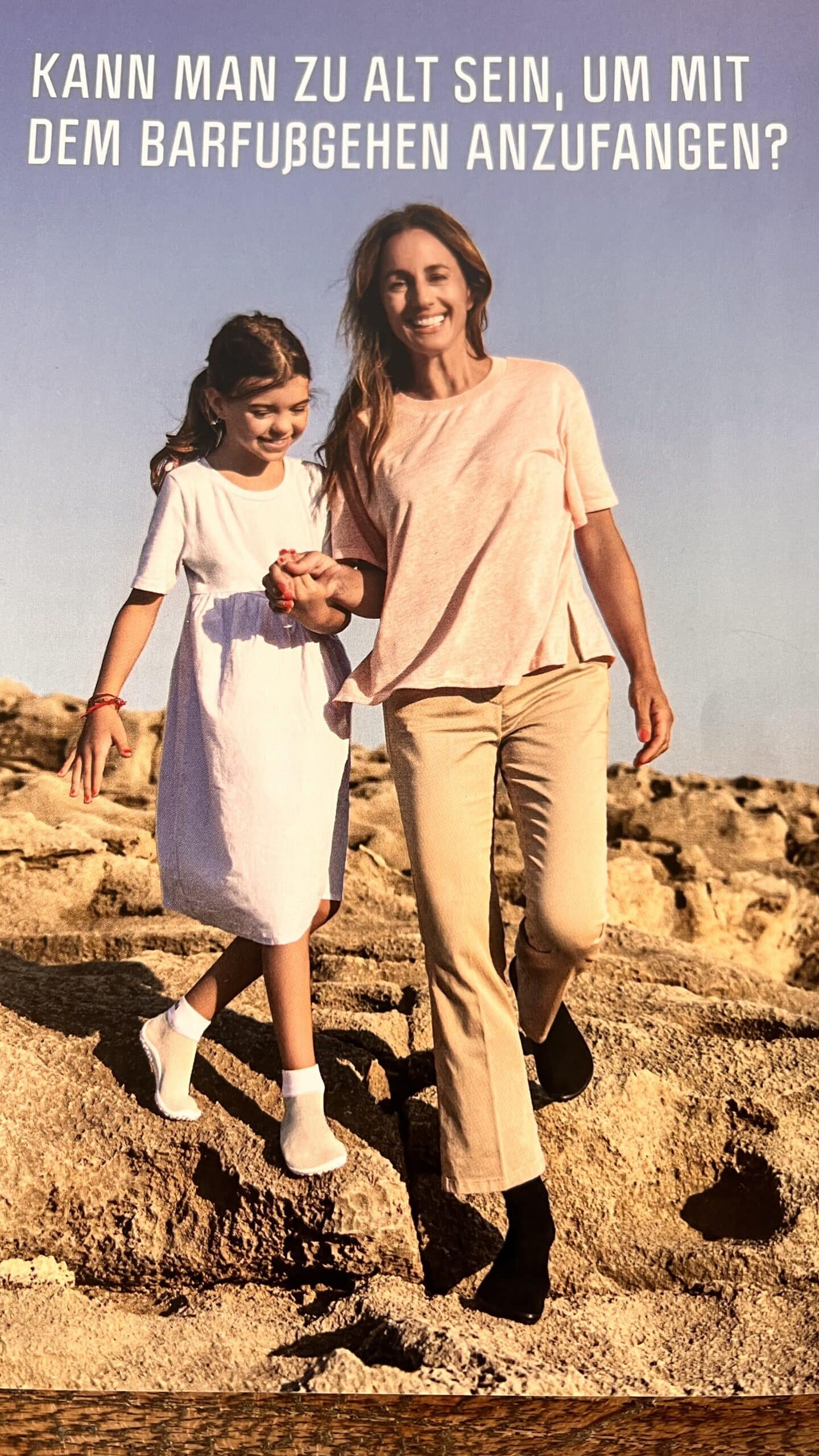 Eine Frau und ein Mädchen laufen barfuß auf steinigem Boden und lächeln unter einem klaren blauen Himmel. Die Frau trägt ein rosa Hemd und beige Hosen, während das Mädchen ein weißes Kleid trägt. Oben im Bild wird deutscher Text angezeigt. © Fotografie Tomas Rodriguez
