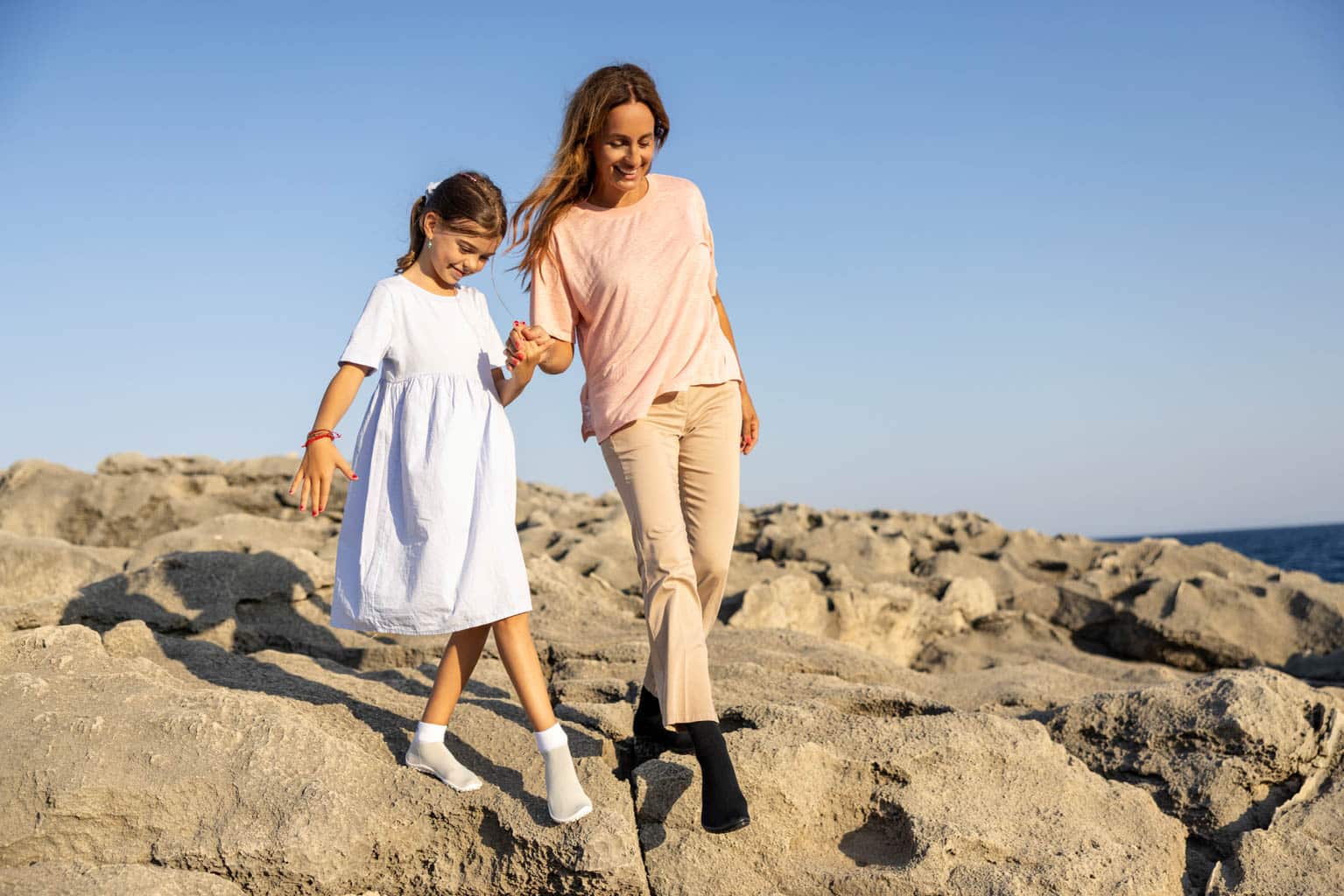 Eine Frau und ein junges Mädchen gehen Händchen haltend über felsiges Gelände am Meer entlang. Die Frau trägt ein pfirsichfarbenes Oberteil und beige Hosen, und das Mädchen trägt ein weißes Kleid. Der Himmel ist klar und blau. © Fotografie Tomas Rodriguez