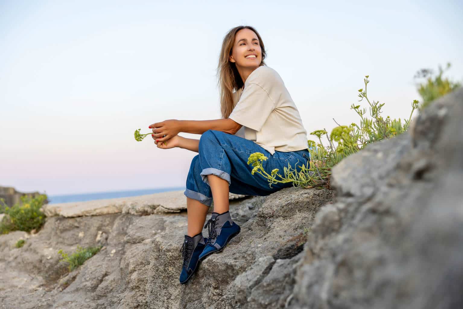 Eine Frau sitzt lächelnd auf einem felsigen Untergrund. Sie trägt ein weißes T-Shirt, Jeans und schwarze Schuhe. Sie ist von kleinen grünen Pflanzen umgeben. Im Hintergrund sind das Meer und der Horizont unter einem klaren Himmel zu sehen. © Fotografie Tomas Rodriguez