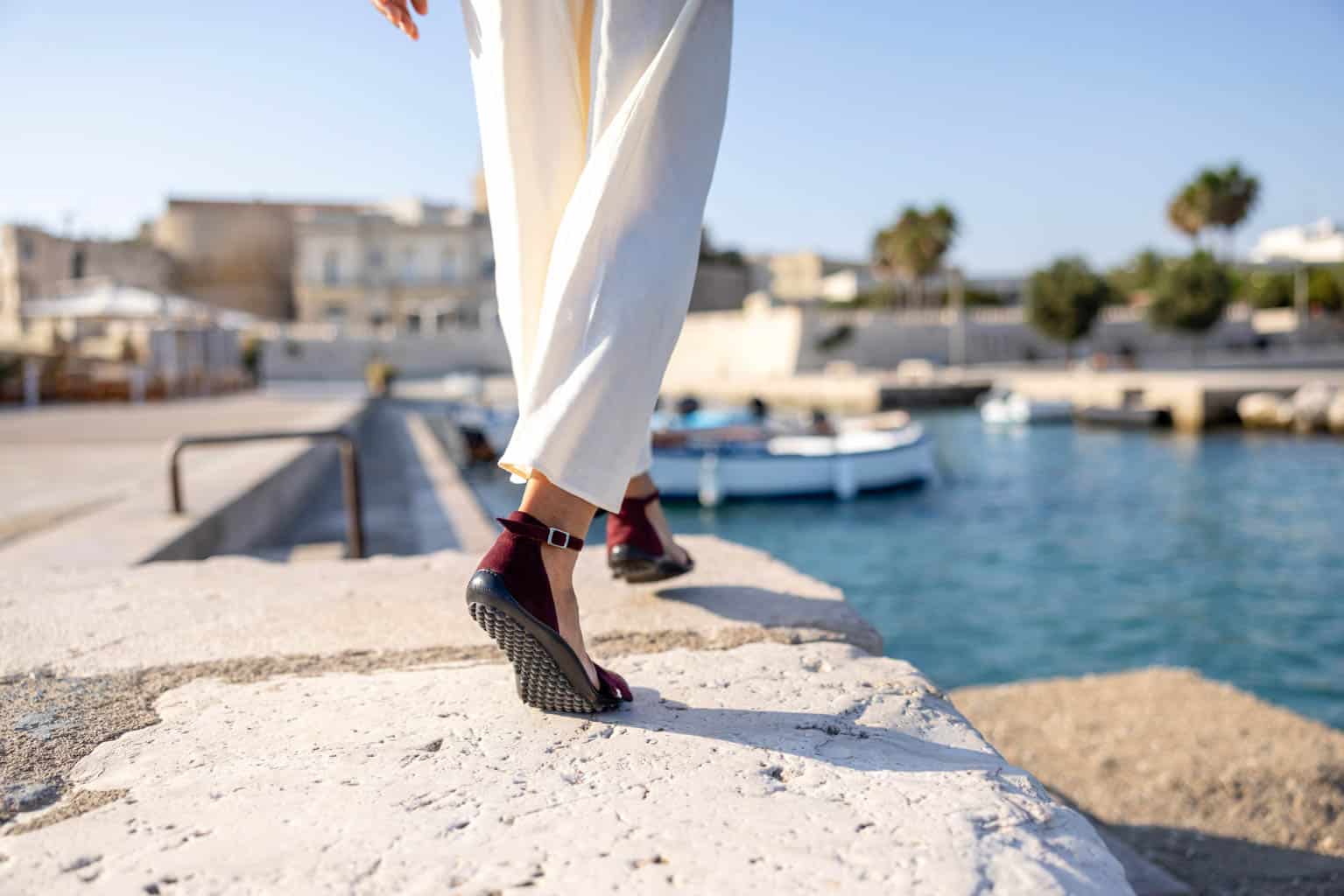 Eine Person in hellen Hosen und kastanienbraunen Sandalen mit Keilabsatz geht auf einer Steinplattform in der Nähe eines Hafens entlang. Im Hintergrund sind Boote auf dem Wasser, Gebäude und Palmen unter einem klaren blauen Himmel zu sehen. © Fotografie Tomas Rodriguez