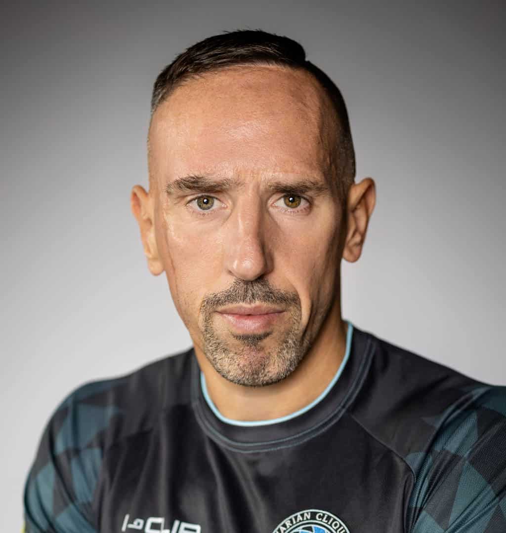 Frank Ribery, Fussballspieler Ein Mann mit kurzen dunklen Haaren und Bart schaut mit neutralem Gesichtsausdruck direkt in die Kamera. Er trägt ein schwarzes Sporttrikot mit einem Text auf hellgrauem Hintergrund. © Fotografie Tomas Rodriguez