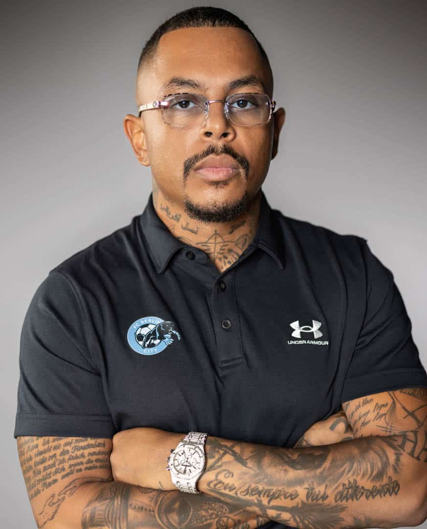 Ein Mann mit Tattoos und Brille trägt ein schwarzes Under Armour-Poloshirt mit Logo darauf. Er steht mit verschränkten Armen da und trägt eine Uhr an seinem Handgelenk. Der Hintergrund ist neutral grau. © Fotografie Tomas Rodriguez
