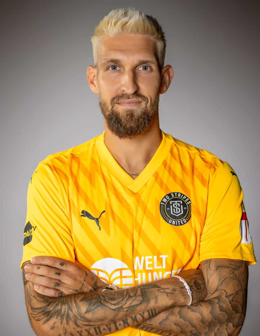 Eine Person mit kurzen blonden Haaren und Bart trägt ein leuchtend gelbes Sporttrikot mit Logos und Texten, darunter „Red Stripes United“ und „Welt Hunger“. Ihre Arme sind verschränkt und zeigen Tattoos. Der Hintergrund ist grau. © Fotografie Tomas Rodriguez