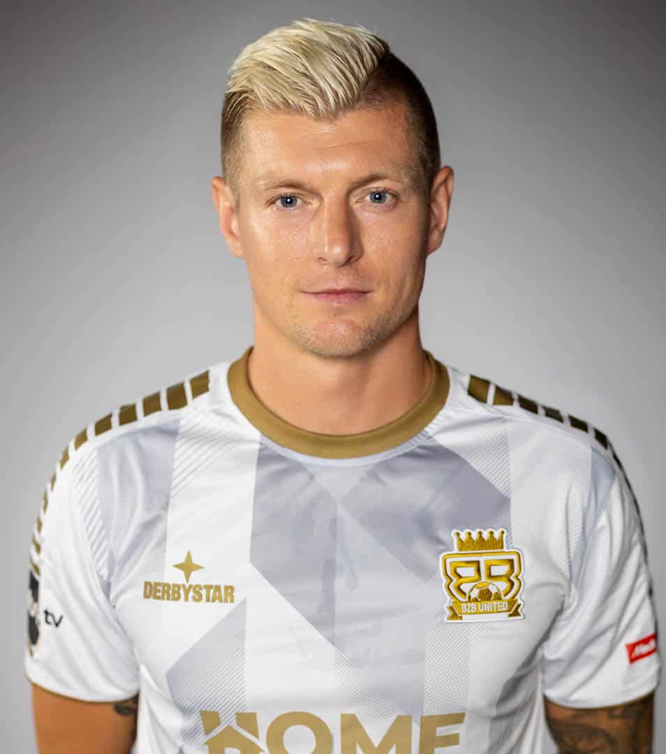 Toni Kroos, Fussballspieler, Eine Person mit kurzen blonden Haaren trägt ein weiß-goldenes Sporttrikot mit Logos und Mustern. Sie steht vor einem neutralen grauen Hintergrund. © Fotografie Tomas Rodriguez