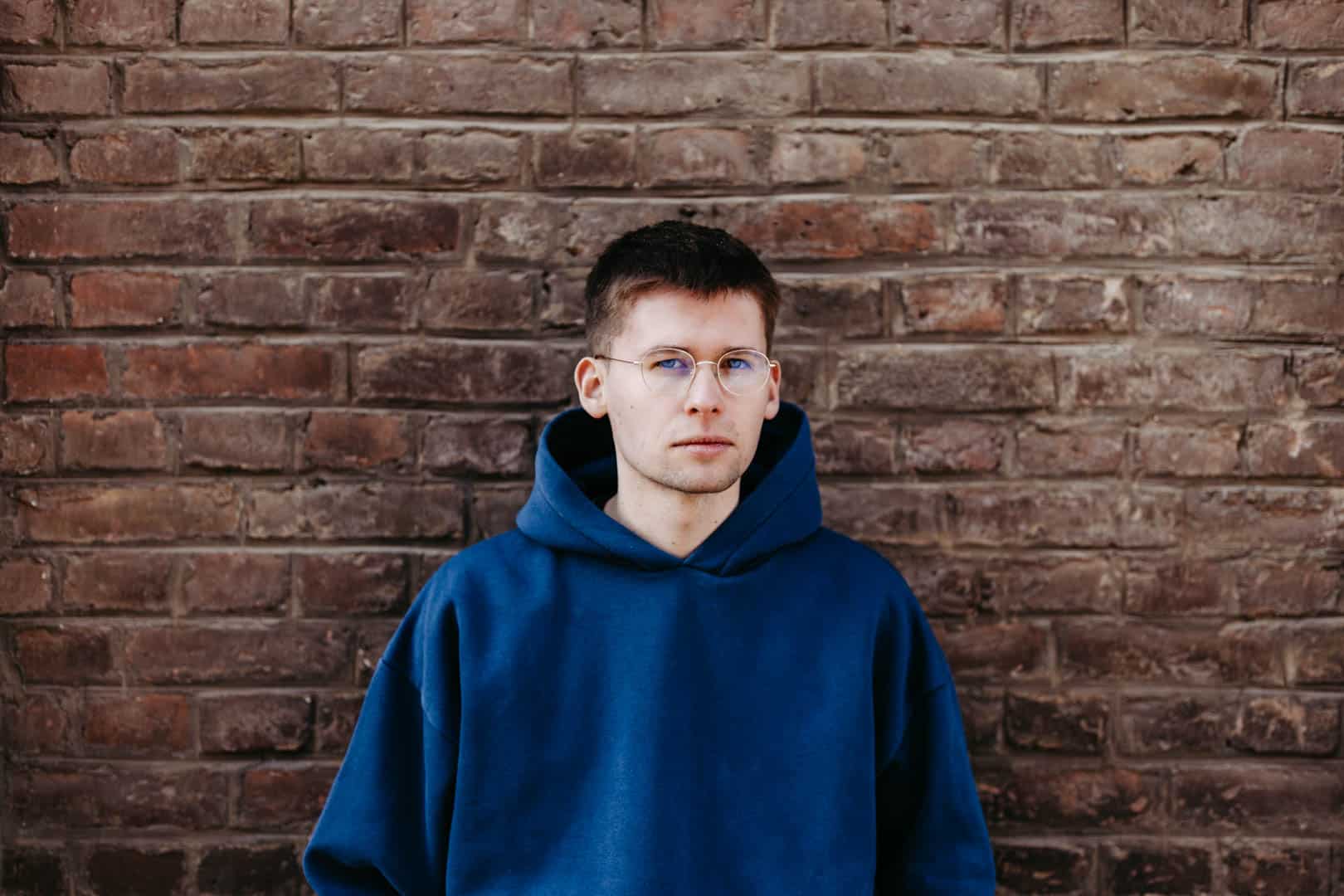 Eine Person mit Brille und blauem Kapuzenpullover steht vor einer Backsteinmauer und blickt direkt in die Kamera. Die Backsteine sind gleichmäßig angeordnet und bilden einen strukturierten Hintergrund. © Fotografie Tomas Rodriguez