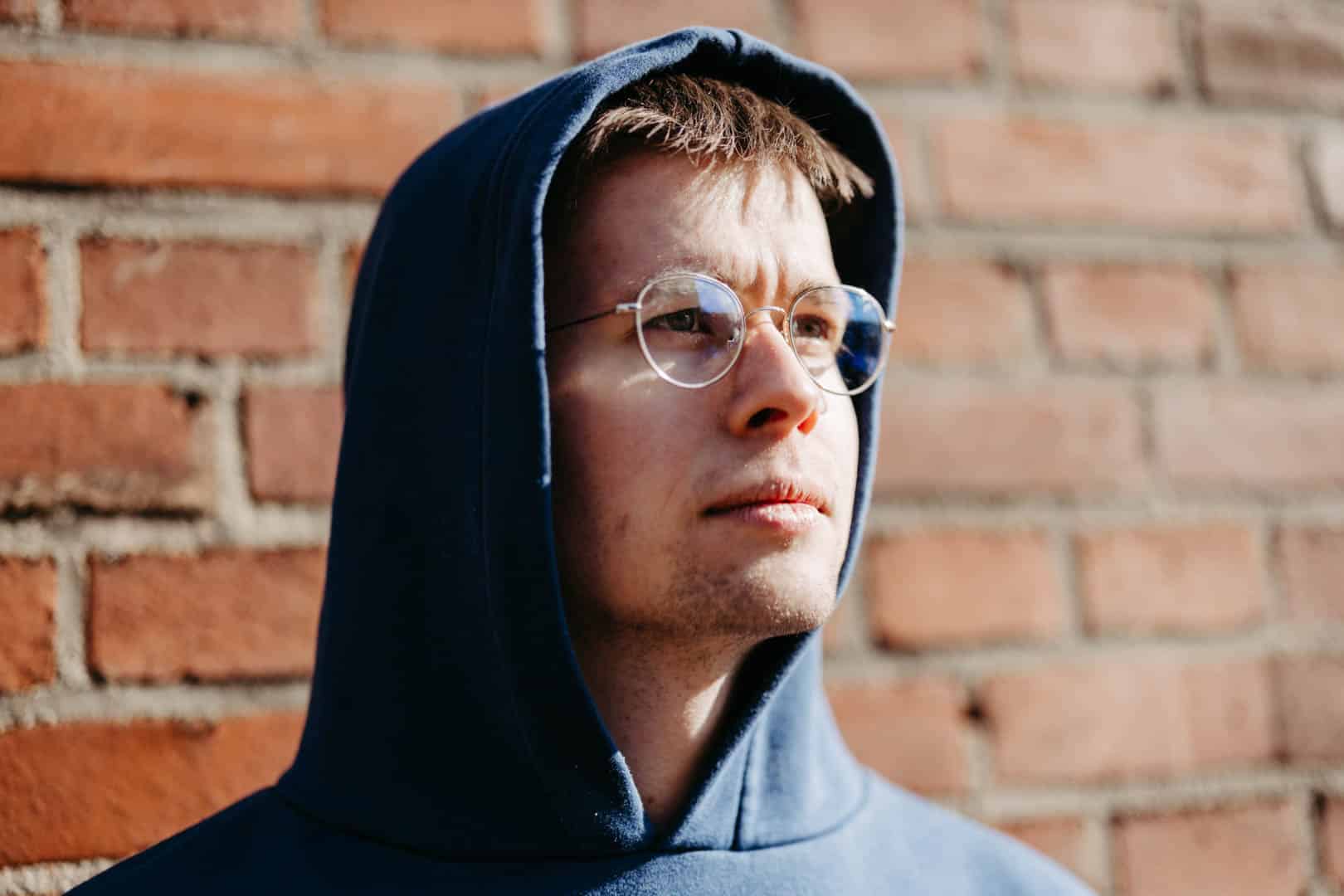 Eine Person mit blauem Kapuzenpullover und Brille steht vor einer Backsteinmauer und blickt nach oben. Das Sonnenlicht wirft einen sanften Schimmer auf ihr Gesicht und hebt die Konturen und Ausdrücke hervor. © Fotografie Tomas Rodriguez