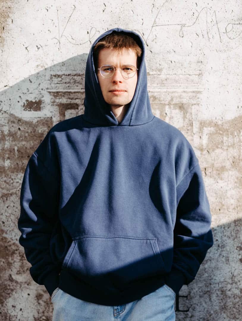 Eine Person mit Brille und blauem Kapuzenpullover steht mit den Händen in den Taschen vor einer strukturierten Betonwand. Schatten auf der Wand lassen Sonnenlicht vermuten. Die Person hat einen neutralen Gesichtsausdruck. © Fotografie Tomas Rodriguez
