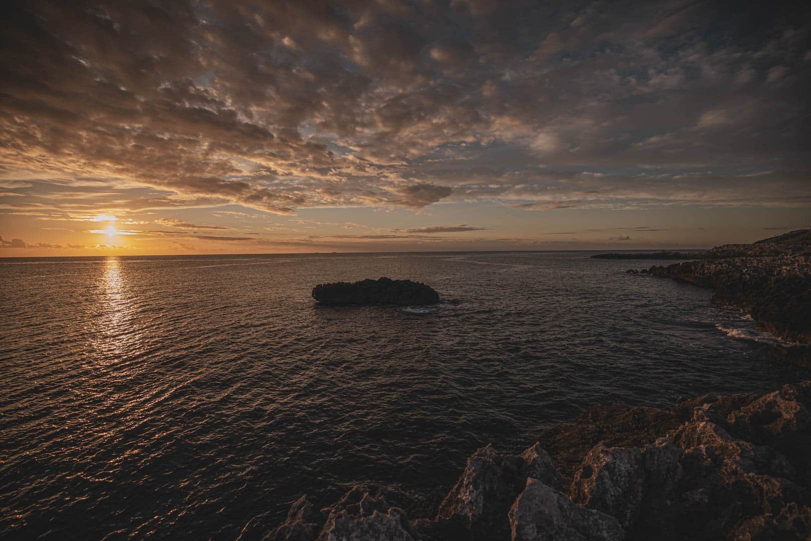 Ein dramatischer Sonnenuntergang über dem Meer mit goldenem Licht, das sich auf dem Wasser spiegelt, einem dunklen Felsen im Vordergrund und einer großen Felsformation in der Mitte des Meeres unter einem Himmel mit dicken Wolken. © Fotografie Tomas Rodriguez