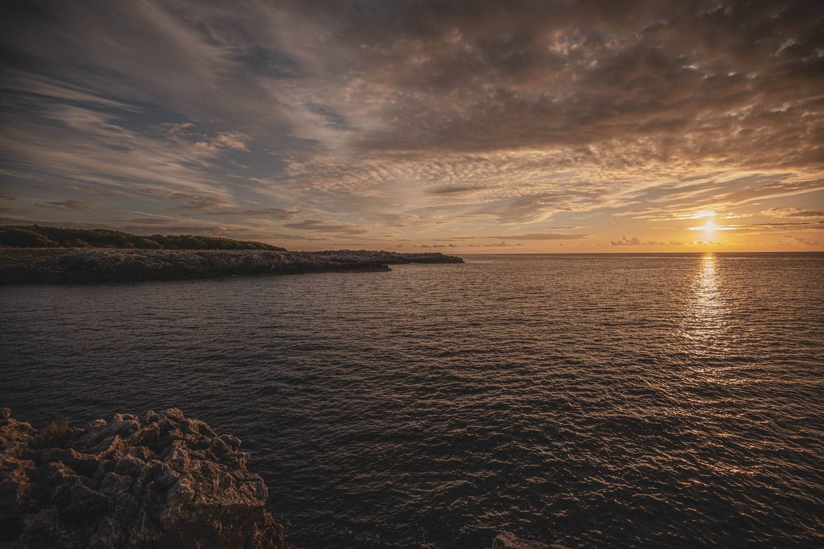 Felsenküste bei Sonnenuntergang, die Sonne steht tief am Horizont und wirft einen goldenen Schein auf das Wasser und den mit dramatischen Wolken gefüllten Himmel. Das Meer erscheint ruhig, und die Küstenlinie reicht bis in die Ferne. © Fotografie Tomas Rodriguez