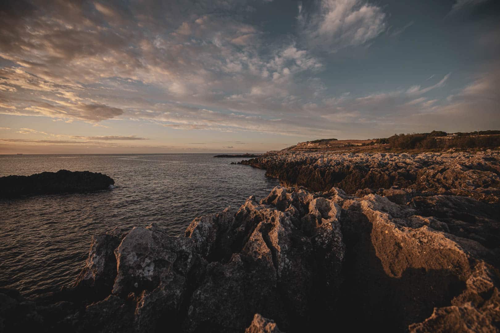 Felsenküste bei Sonnenuntergang mit dramatischen Wolken am Himmel, schroffen Felsen im Vordergrund und ruhiger See am Horizont. Das warme Sonnenlicht hebt die Texturen der Felsen und Wolken hervor. © Fotografie Tomas Rodriguez