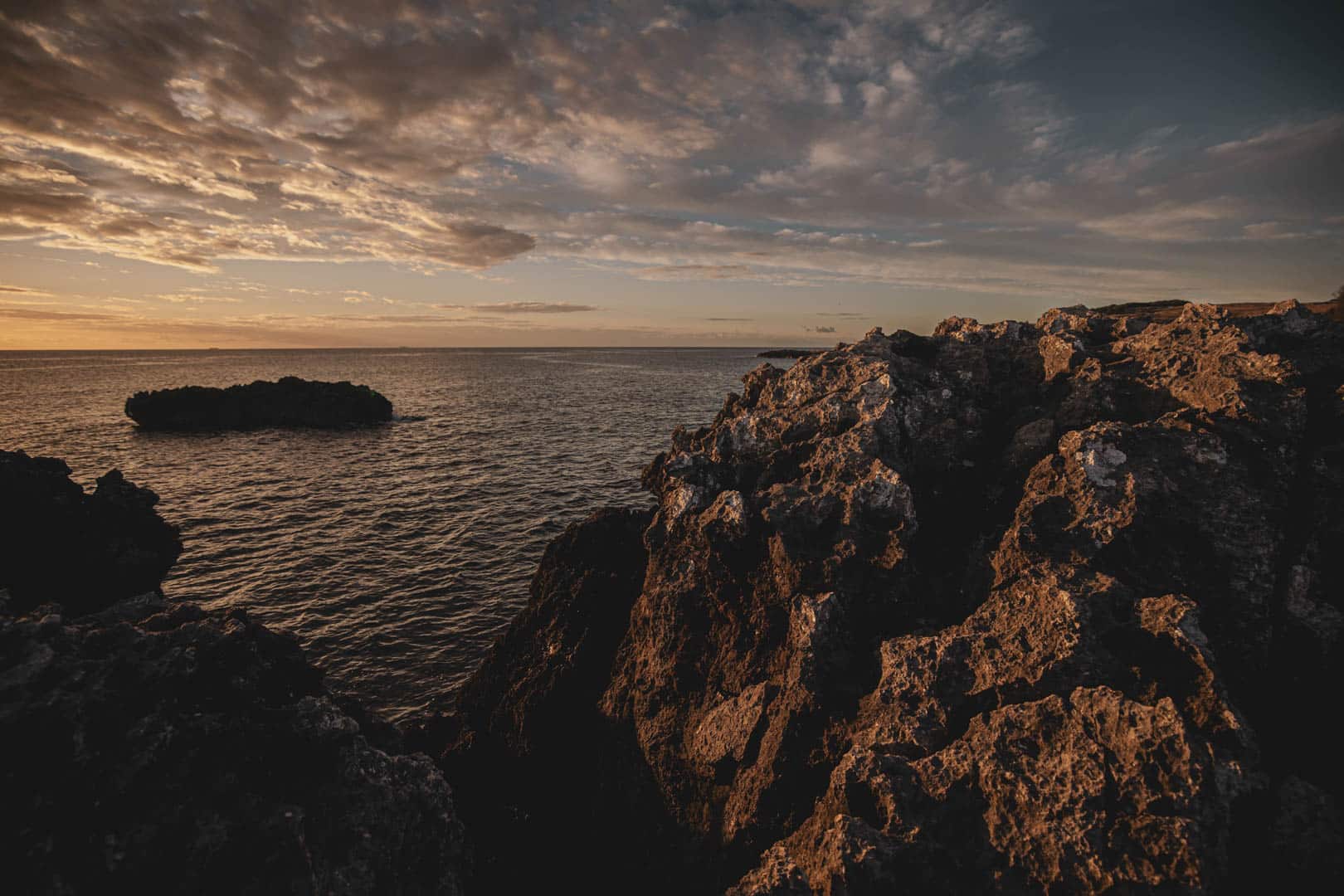Zerklüftete Felsklippen überblicken das ruhige Meer bei Sonnenuntergang, mit dramatischen Wolken am Himmel und warmem Licht, das Schatten auf die Felsen wirft. © Fotografie Tomas Rodriguez