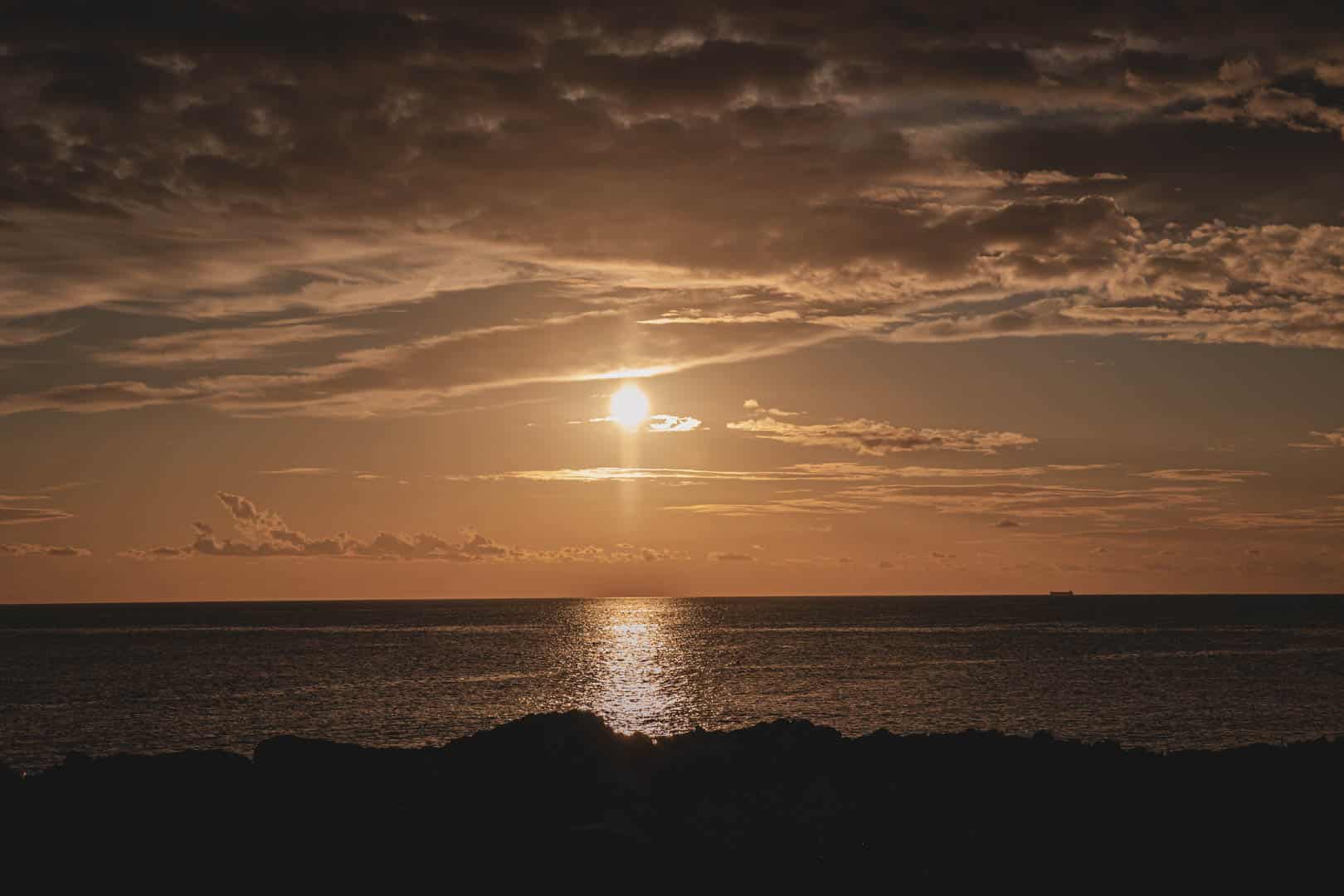 Goldener Sonnenuntergang über dem Meer mit dramatischen Wolken, Sonnenlicht, das sich auf dem Wasser spiegelt, und dunklen Felssilhouetten im Vordergrund. © Fotografie Tomas Rodriguez