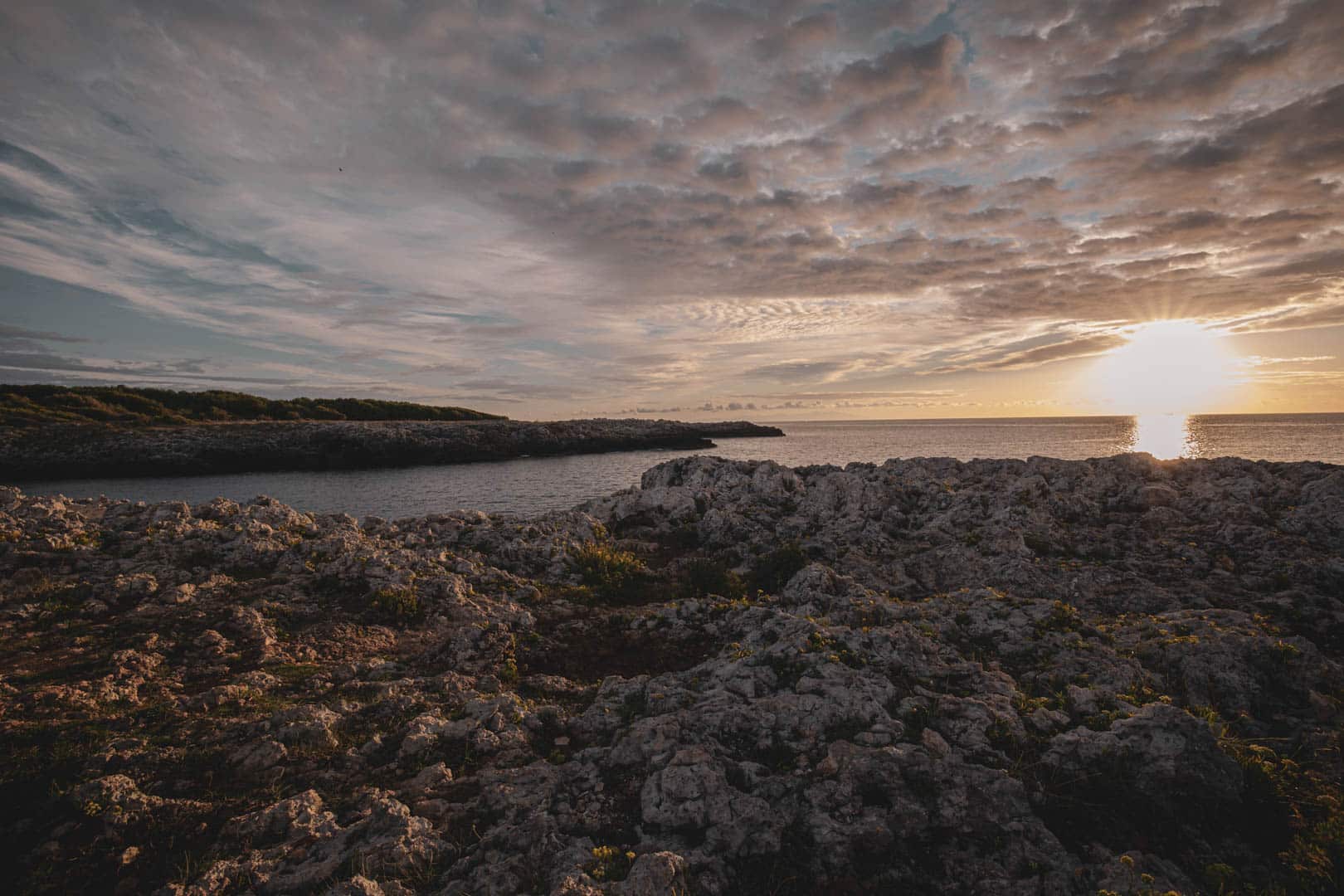 Felsenküste bei Sonnenuntergang, die Sonne steht tief am Horizont und wirft warmes Licht auf die Felsen und das Wasser. Der Himmel ist teilweise bewölkt, was für dramatische Lichtverhältnisse und Spiegelungen auf dem Meer sorgt. © Fotografie Tomas Rodriguez