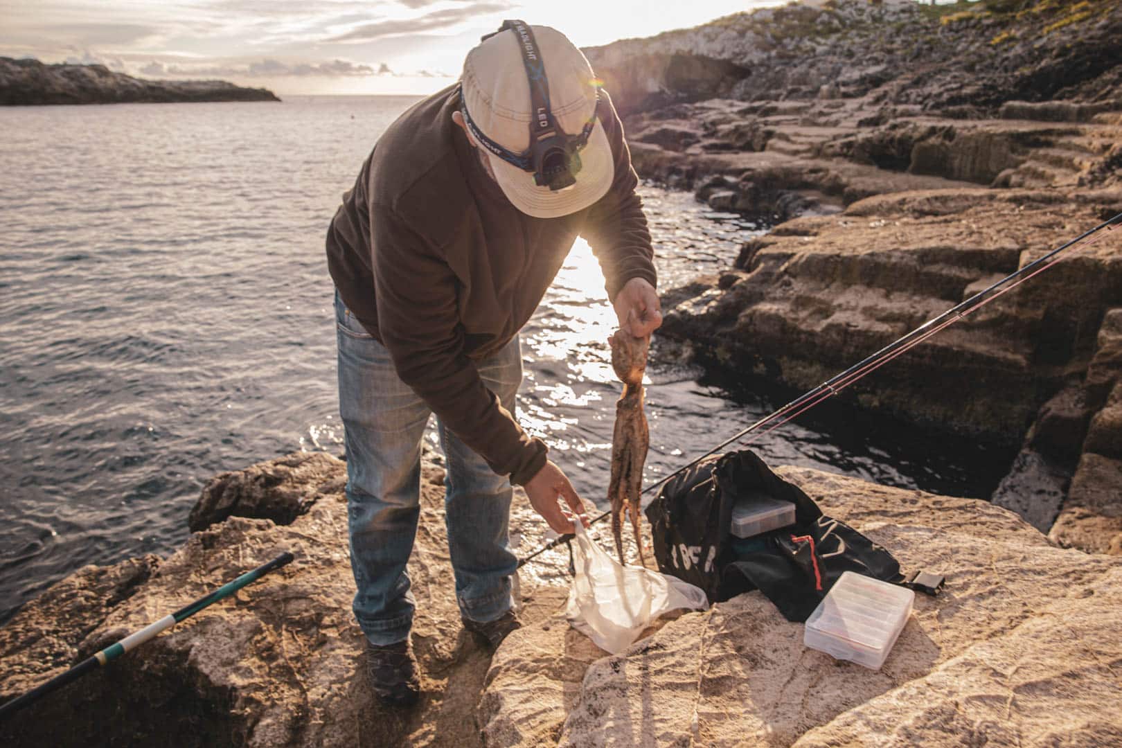 Eine Person mit einer Stirnlampe steht am felsigen Ufer des Meeres und hält einen frisch gefangenen Tintenfisch neben einer Angelrute und einem Angelkasten, um den Fang bei Sonnenuntergang zu erlegen. © Fotografie Tomas Rodriguez