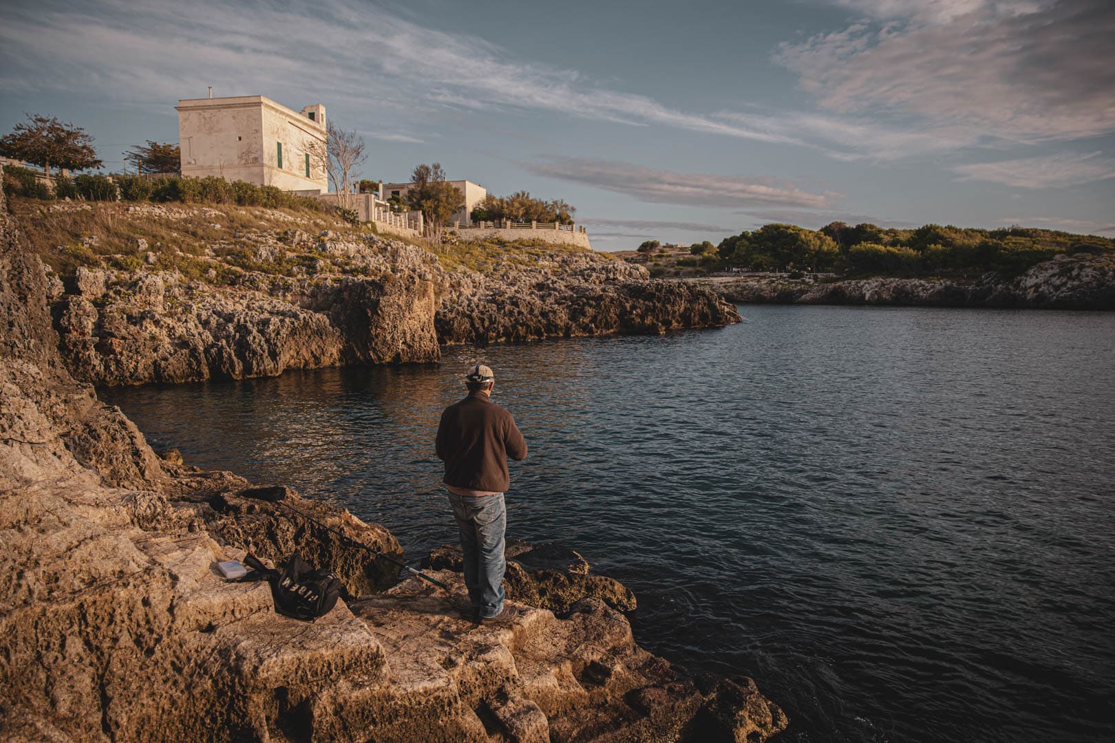 Eine Person steht am felsigen Ufer und angelt im ruhigen Wasser, mit schroffen Klippen und einem großen Gebäude auf einem Hügel im Hintergrund unter einem teilweise bewölkten Himmel. © Fotografie Tomas Rodriguez