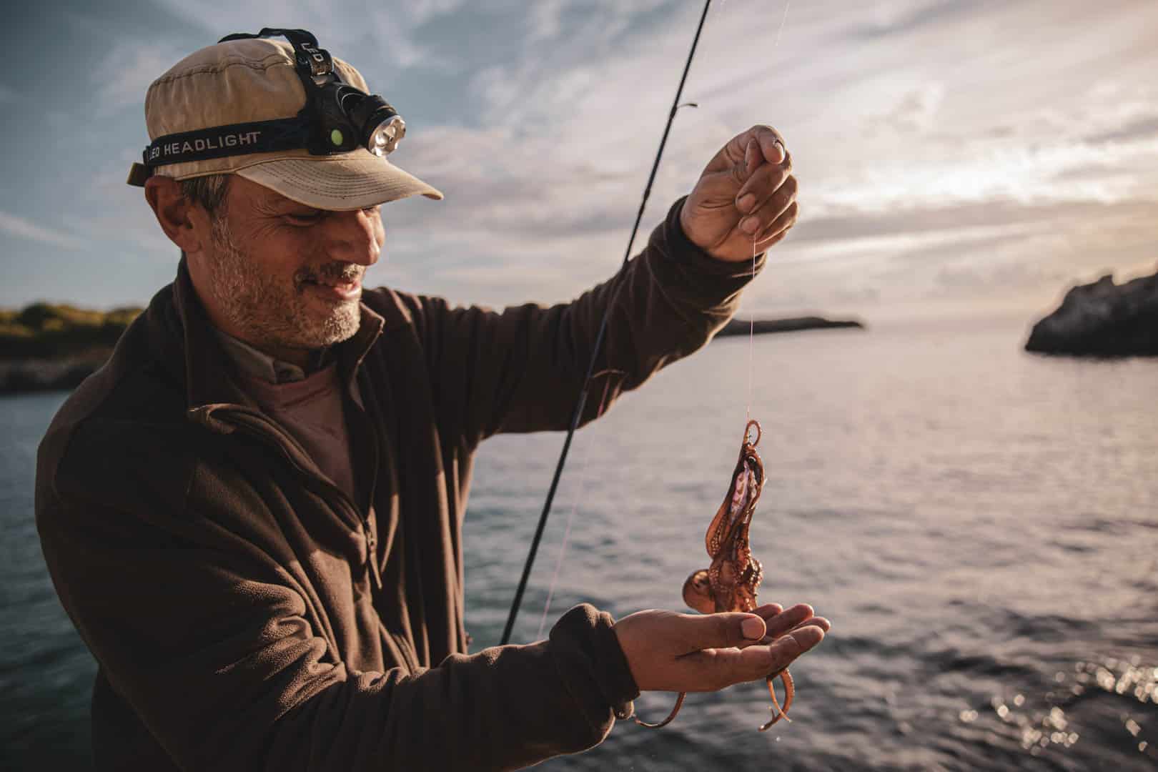 Eine Person mit Mütze und Stirnlampe lächelt, während sie einen kleinen Tintenfisch in der Hand hält, der an einer Angelschnur am Meer gefangen ist. Im Hintergrund sind felsige Küsten und ein bewölkter Himmel zu sehen. © Fotografie Tomas Rodriguez