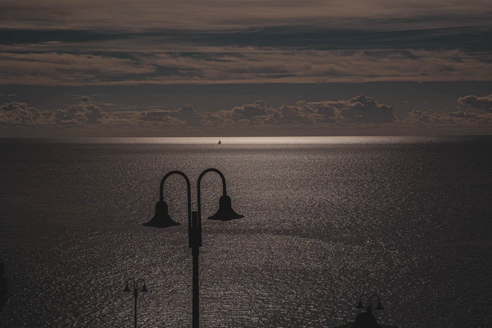 Eine dunkle, stimmungsvolle Meereslandschaft in der Abenddämmerung mit schimmerndem Wasser, silhouettierten Straßenlaternen im Vordergrund und einem einzelnen, weit entfernten Segelboot am Horizont unter einem bewölkten Himmel. © Fotografie Tomas Rodriguez