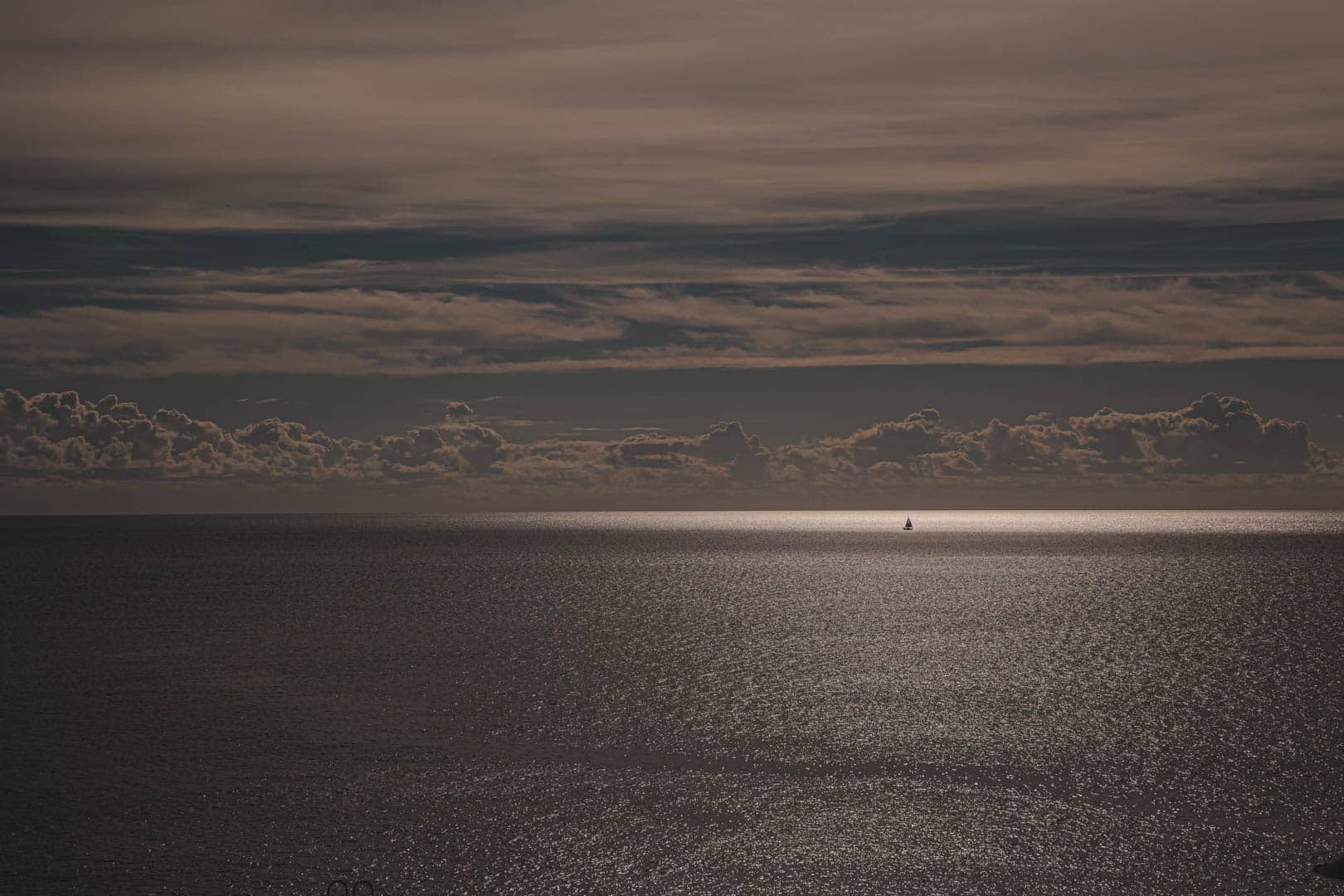 Ein kleines Segelboot ist auf einem schimmernden, sonnenbeschienenen Meer unter einem dramatischen Himmel mit dunklen Wolken und Sonnenflecken zu sehen, die sich auf dem Wasser spiegeln. © Fotografie Tomas Rodriguez