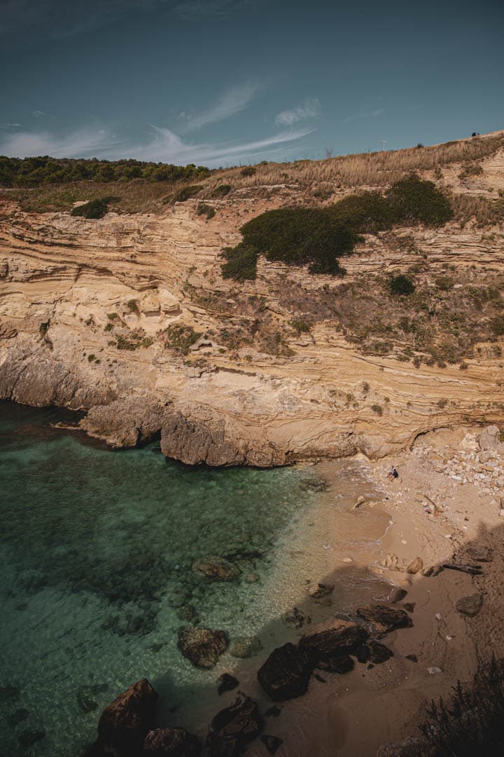 Ein abgelegener Sandstrand mit klarem, türkisfarbenem Wasser und felsigen Klippen, gesäumt von Sträuchern und Gras unter einem teilweise bewölkten Himmel. © Fotografie Tomas Rodriguez