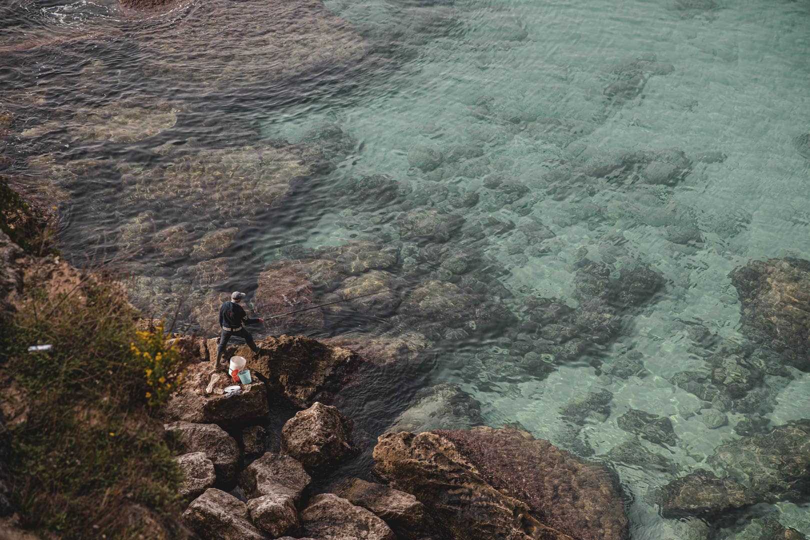 Eine Person steht am felsigen Ufer und angelt mit einer Angelrute, umgeben von klarem türkisfarbenem Wasser und untergetauchten Felsen; in der Nähe stehen ein Eimer und Zubehör. Die Szene ist von oben betrachtet. © Fotografie Tomas Rodriguez