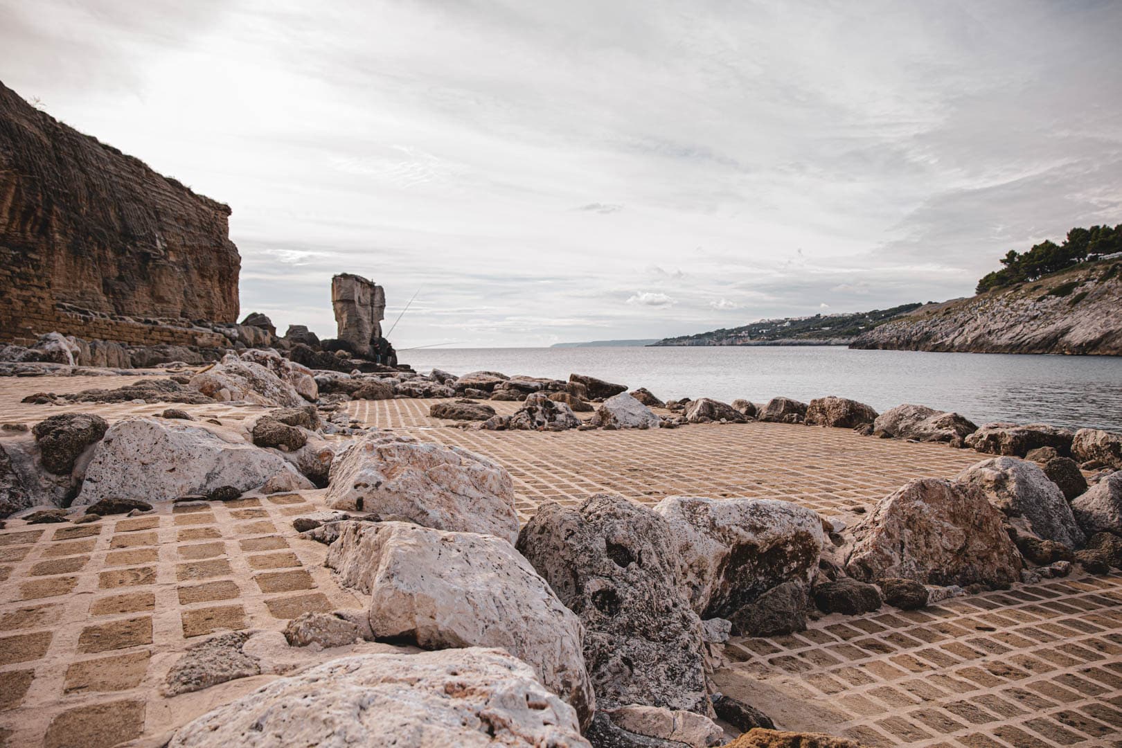 Küstenszene mit großen Felsen und Steingittern am Meer, einer hohen Felsformation in Küstennähe und entfernten Klippen mit etwas Grün unter einem bewölkten Himmel. © Fotografie Tomas Rodriguez