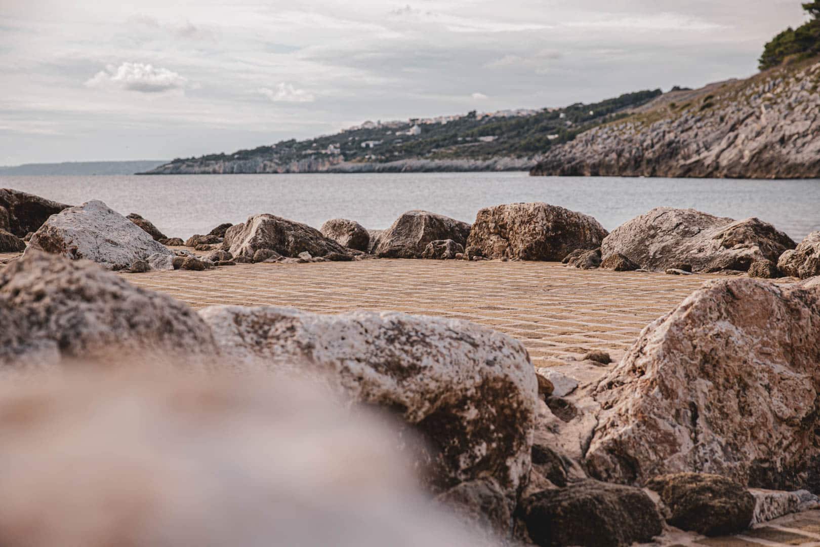 Von einer mit Steinen gepflasterten Plattform, die von großen Felsen umgeben ist, blickt man auf ein ruhiges Meer, mit felsigen Hügeln und einem wolkenverhangenen Himmel im Hintergrund. © Fotografie Tomas Rodriguez