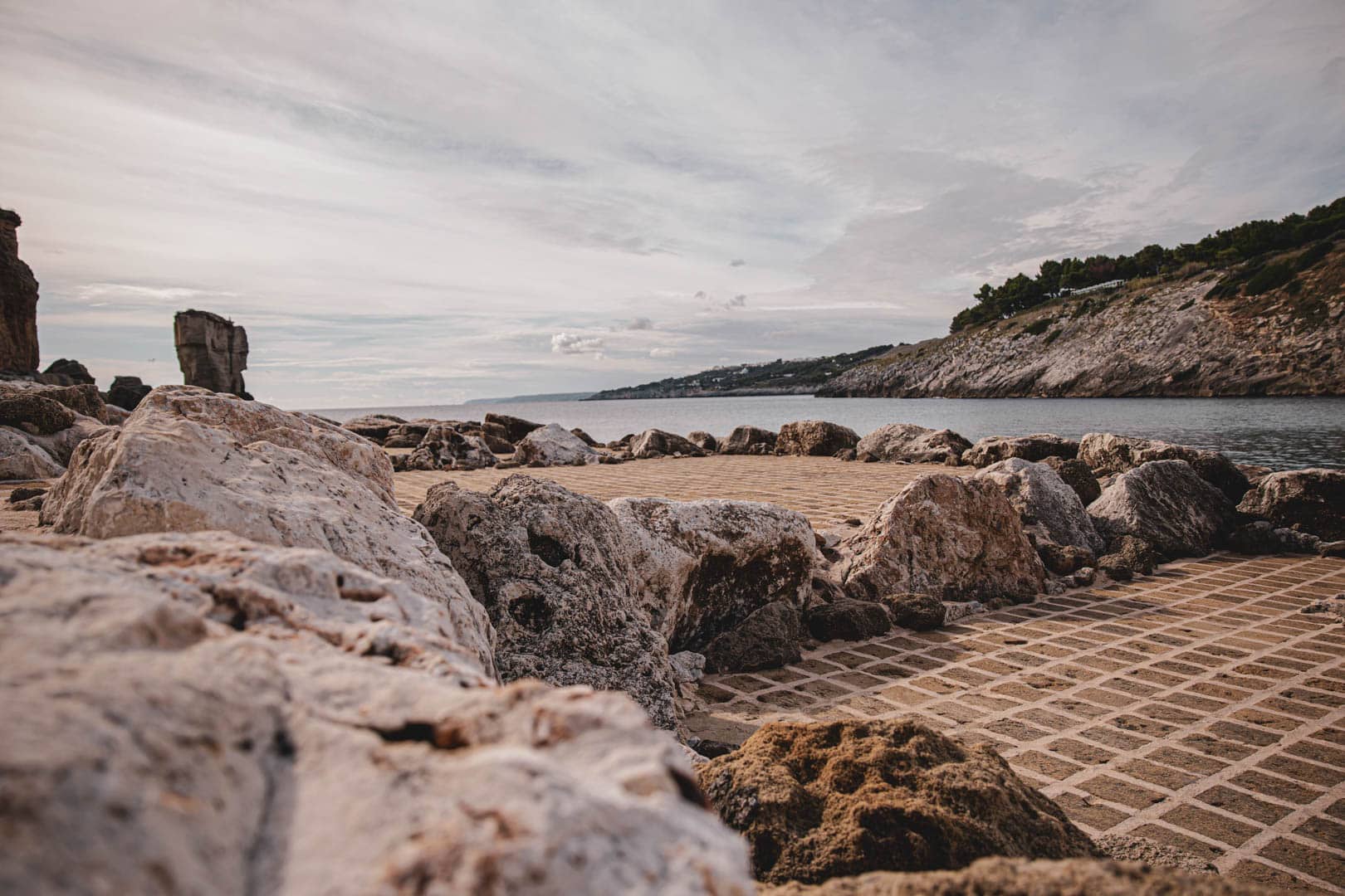 Große Felsen säumen eine gemusterte Steinplattform am Meer, mit felsigen Hängen und entferntem Grün unter einem bewölkten Himmel. © Fotografie Tomas Rodriguez