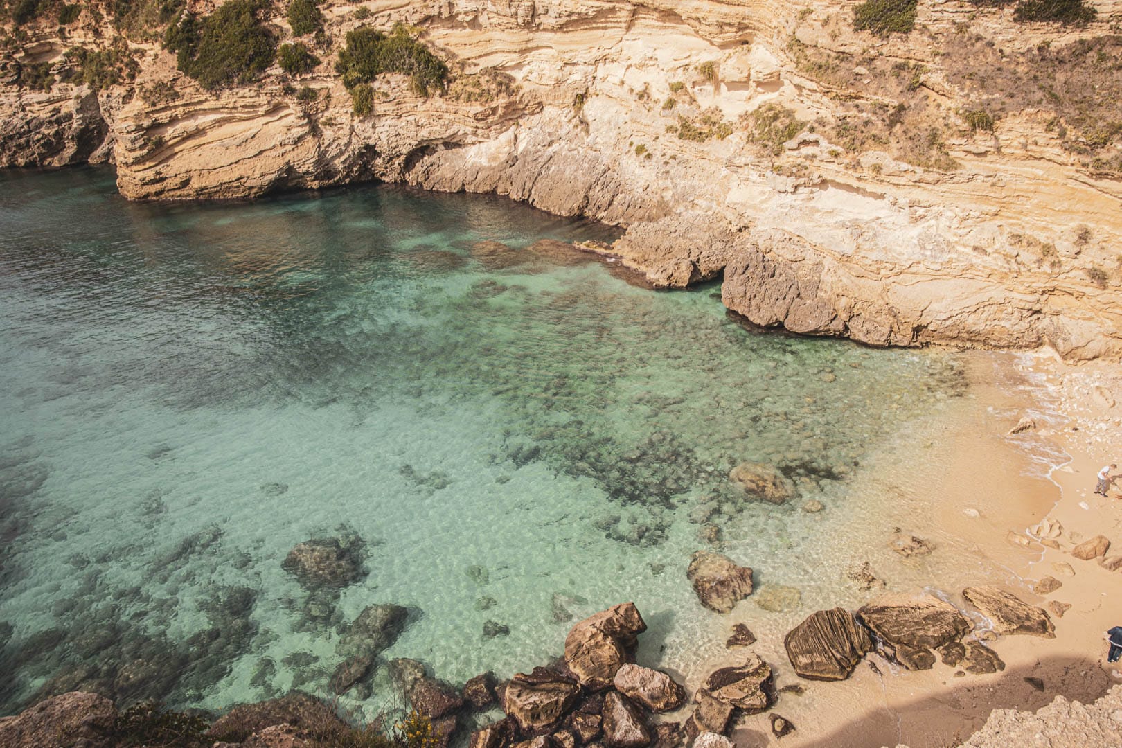 Klares türkisfarbenes Wasser plätschert sanft gegen einen kleinen Sandstrand, der von zerklüfteten, geschichteten Klippen und verstreuten Felsen umgeben ist und eine abgeschiedene und ruhige Küstenszene schafft. © Fotografie Tomas Rodriguez