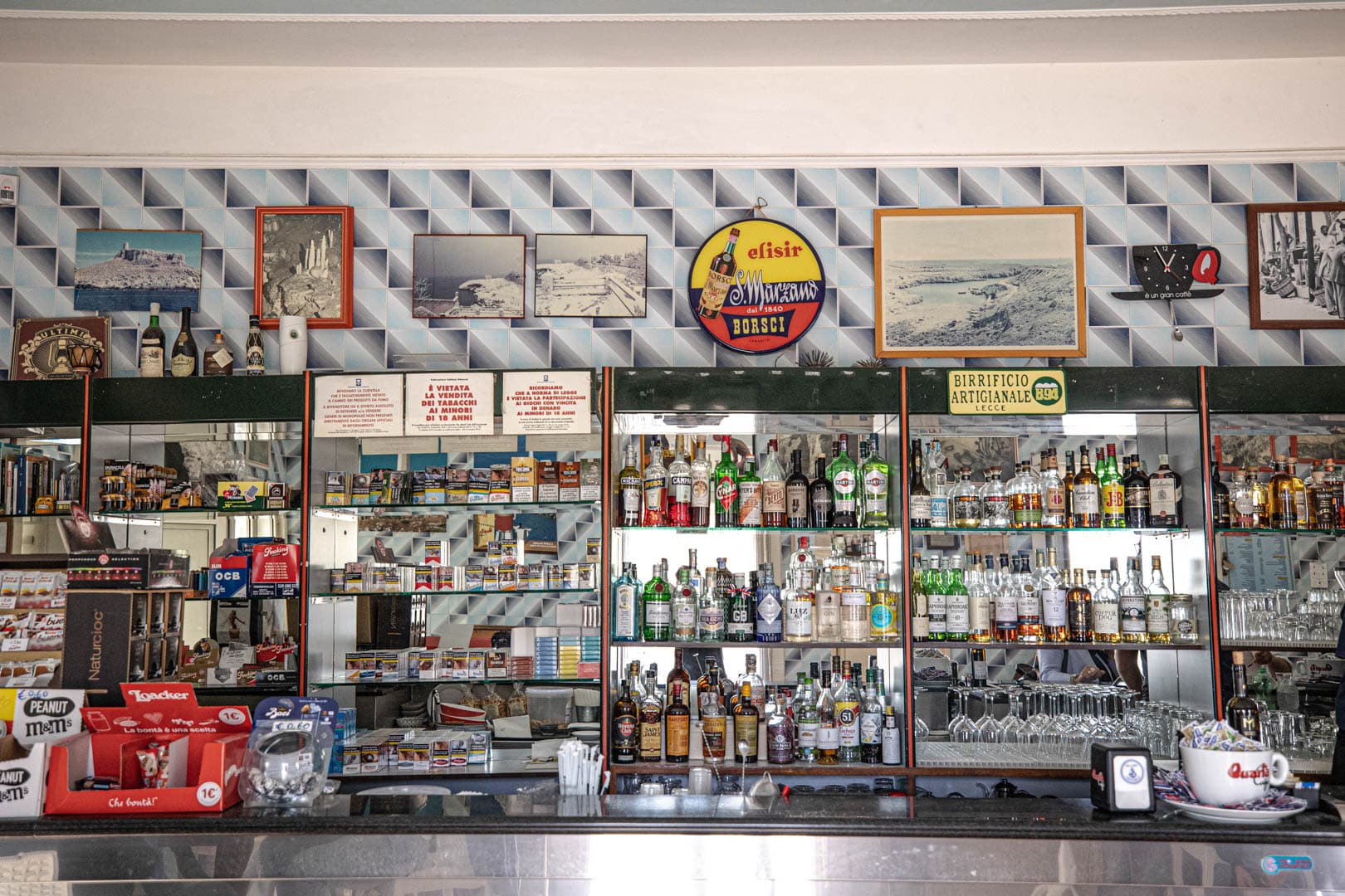 Ein Bartresen mit verschiedenen Alkoholflaschen, Gläsern und Kaffeemaschinen; dahinter spiegeln Spiegel den Raum, und die Wand über den Regalen ist mit alten Fotos und Postern geschmückt. © Fotografie Tomas Rodriguez