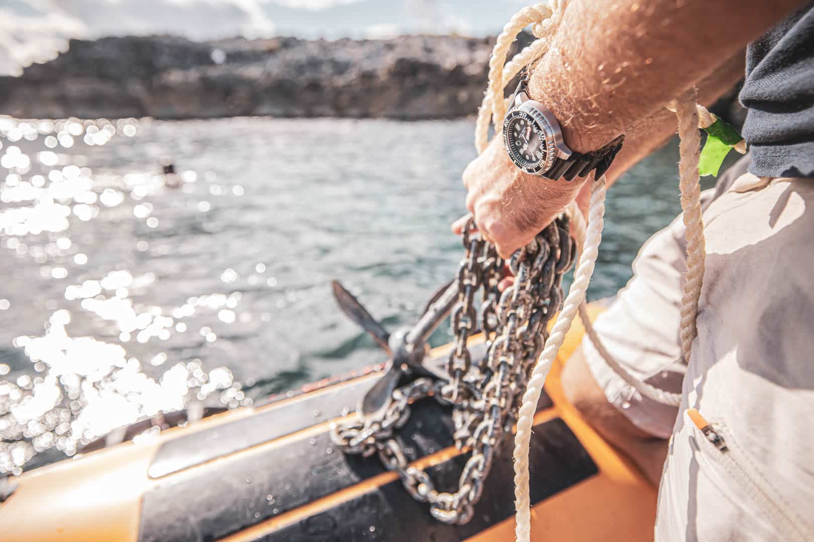 Nahaufnahme einer Person, die ein Seil und eine Metallkette hält, die an einem Anker auf einem Schlauchboot befestigt sind, mit Wasser und einer felsigen Uferlinie im Hintergrund. Die Person trägt eine Armbanduhr. © Fotografie Tomas Rodriguez