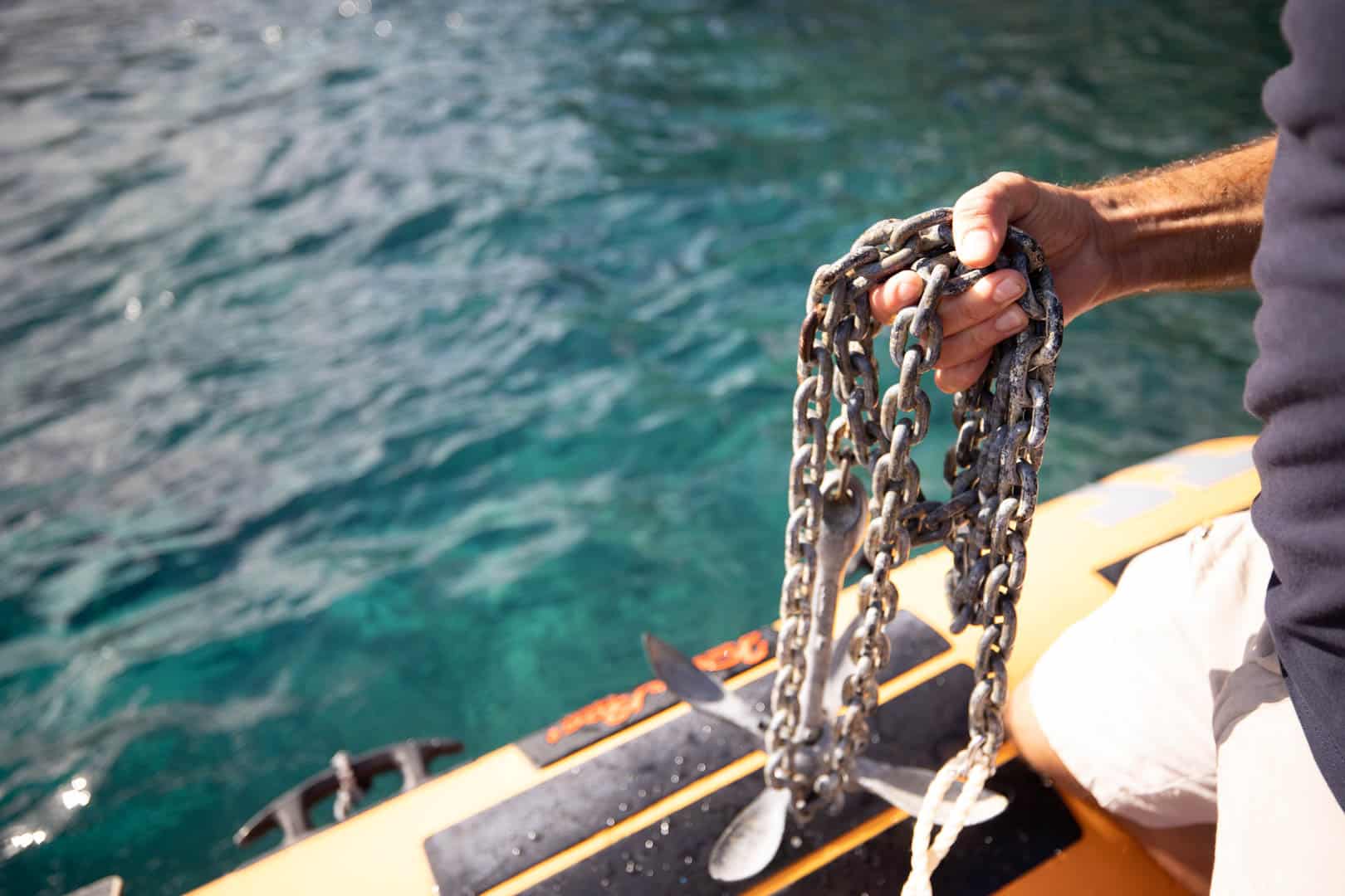 Eine Person hält einen Anker mit einer Kette über den Rand eines gelben Schlauchboots und bereitet sich darauf vor, es in das klare blaue Wasser hinabzulassen. © Fotografie Tomas Rodriguez
