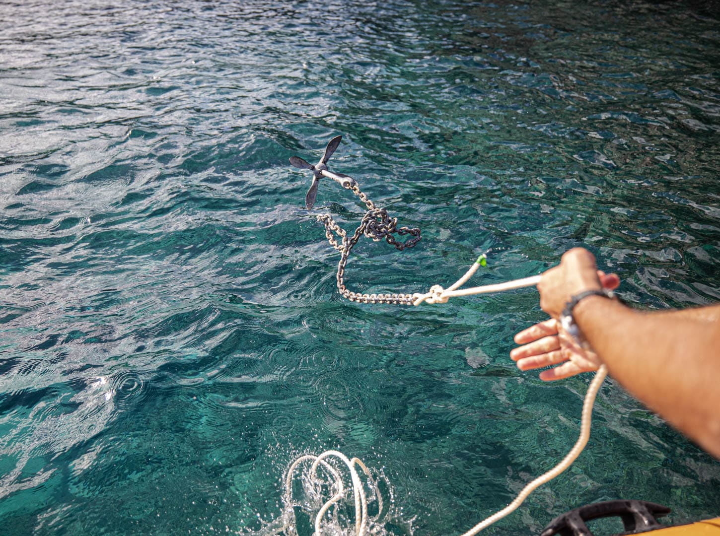 Die Hand einer Person wirft einen Metallanker, der an einer Kette und einem Seil befestigt ist, von einem Boot aus in klares, blaugrünes Wasser, in dem Wellen auf der Wasseroberfläche sichtbar sind. © Fotografie Tomas Rodriguez