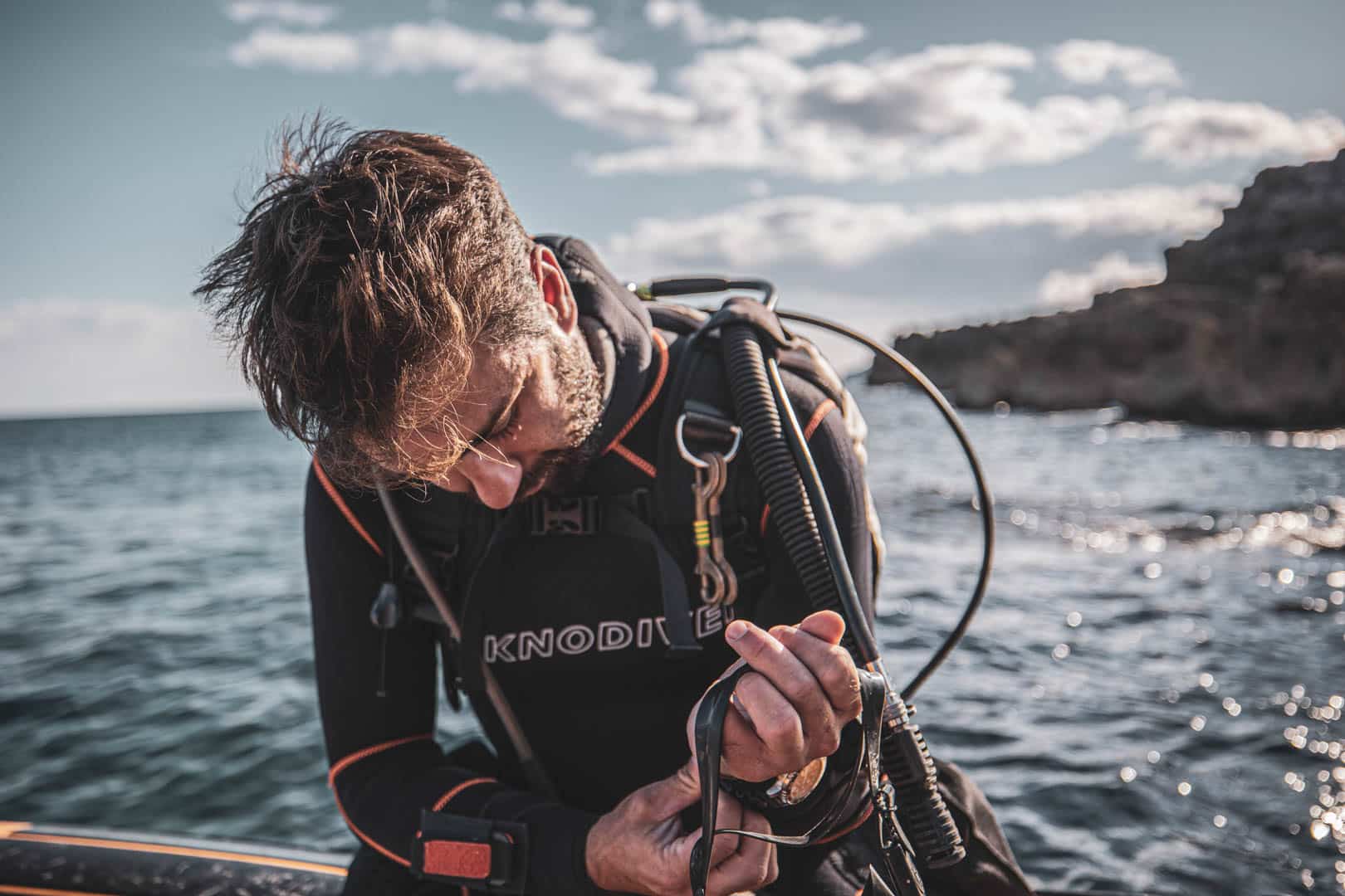 Ein Taucher im Neoprenanzug kniet auf einem Boot am Meer und richtet seine Tauchausrüstung im hellen Sonnenlicht aus, mit felsigen Klippen und blauem Wasser im Hintergrund. © Fotografie Tomas Rodriguez
