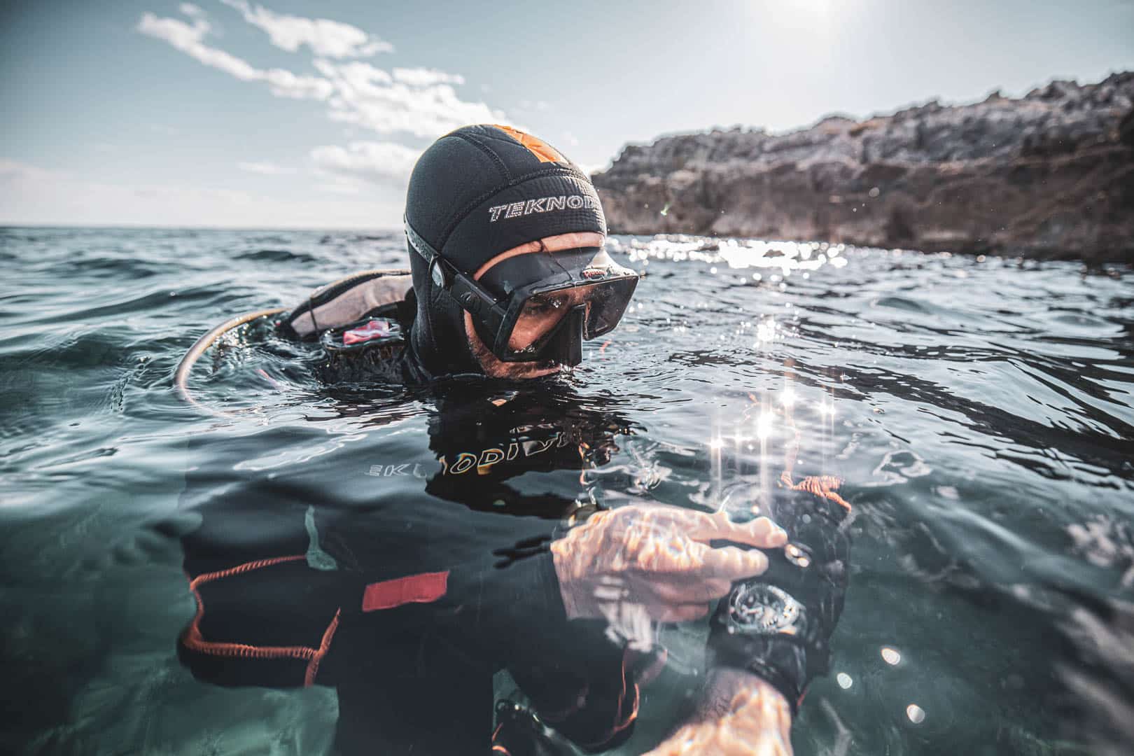 Ein Taucher mit Neoprenanzug und Maske schwimmt im Meer in der Nähe des felsigen Ufers und blickt auf eine Taucheruhr an seinem Handgelenk, während sich das Sonnenlicht auf der Wasseroberfläche spiegelt. © Fotografie Tomas Rodriguez
