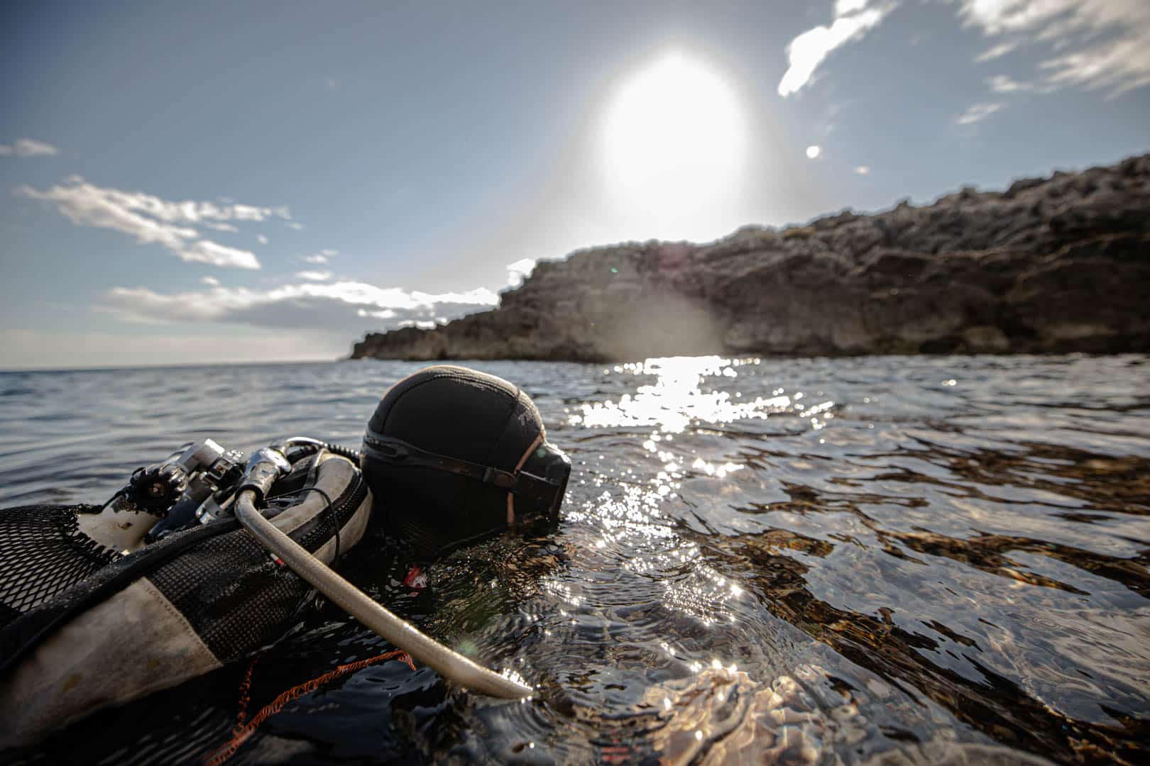 Ein Taucher mit Neoprenanzug und Maske schwimmt im flachen, sonnenbeschienenen Wasser in der Nähe einer felsigen Küste, während die Sonne tief steht und Wolken über ihm hängen. © Fotografie Tomas Rodriguez