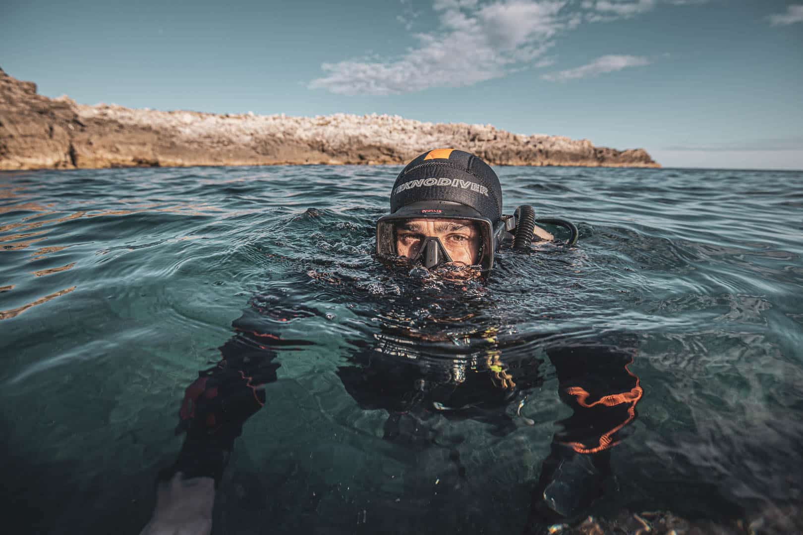 Ein Taucher in Neoprenanzug und Maske schwimmt an der Oberfläche des klaren blauen Wassers in der Nähe der felsigen Küste unter einem teilweise bewölkten Himmel. © Fotografie Tomas Rodriguez