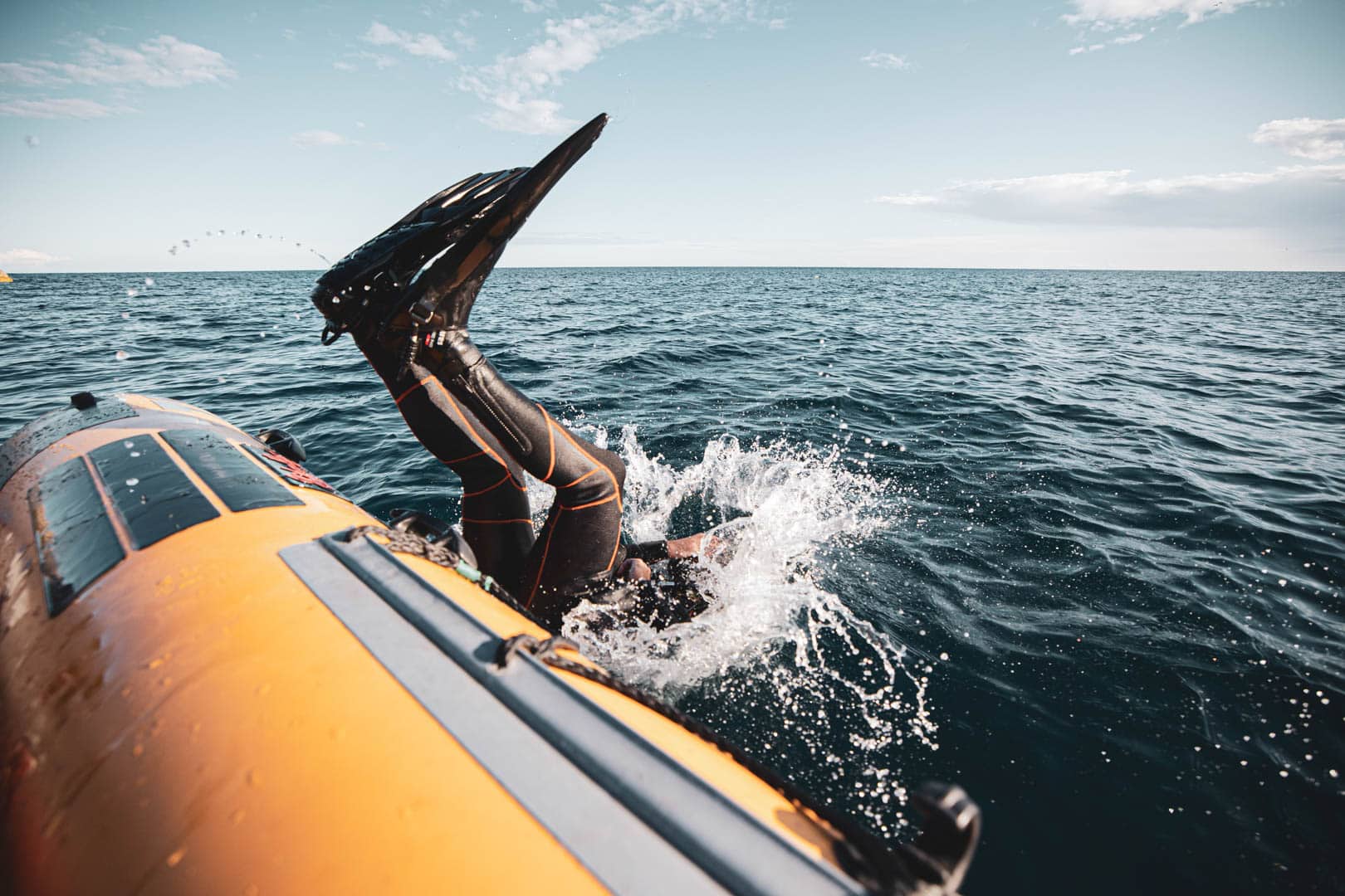 Eine Person, die einen Neoprenanzug und Flossen trägt, springt von der Seite eines orangefarbenen Schlauchboots ins Meer, mit dem Himmel und dem Horizont im Hintergrund. © Fotografie Tomas Rodriguez