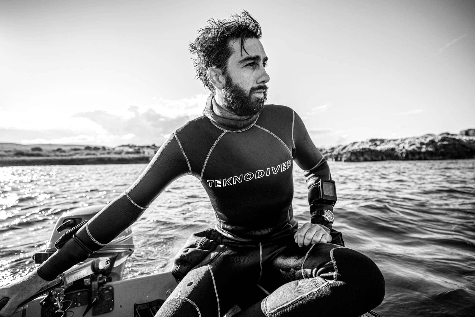Ein Mann in einem Neoprenanzug mit der Aufschrift "TEKNODIVE" sitzt auf einem Boot am Wasser und schaut zur Seite. Er trägt eine Taucheruhr und eine Tauchausrüstung. Im Hintergrund sind felsige Ufer und ruhiges Wasser zu sehen. Das Bild ist in schwarz-weiß gehalten. © Fotografie Tomas Rodriguez