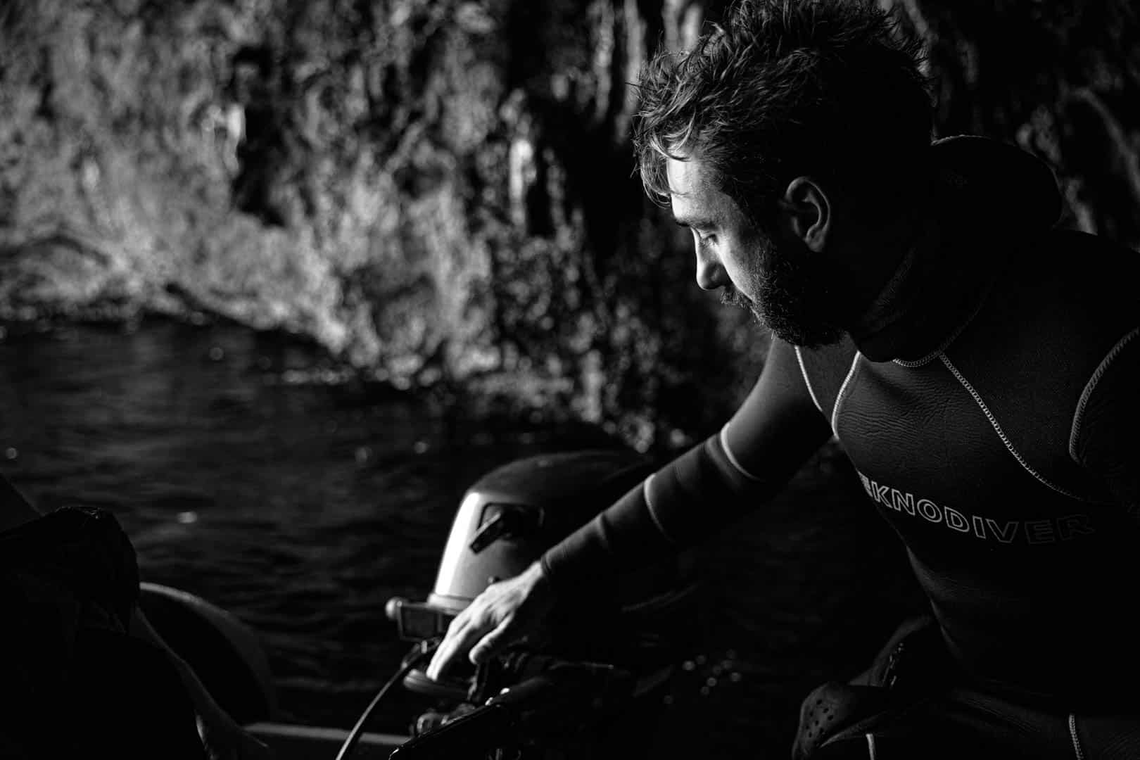 Ein Mann in einem Neoprenanzug steht in der Nähe eines felsigen Ufers, schaut nach unten und bedient die Ausrüstung eines Bootes. Das Bild ist in Schwarz-Weiß gehalten, wobei die dramatische Beleuchtung seinen konzentrierten Ausdruck hervorhebt. © Fotografie Tomas Rodriguez